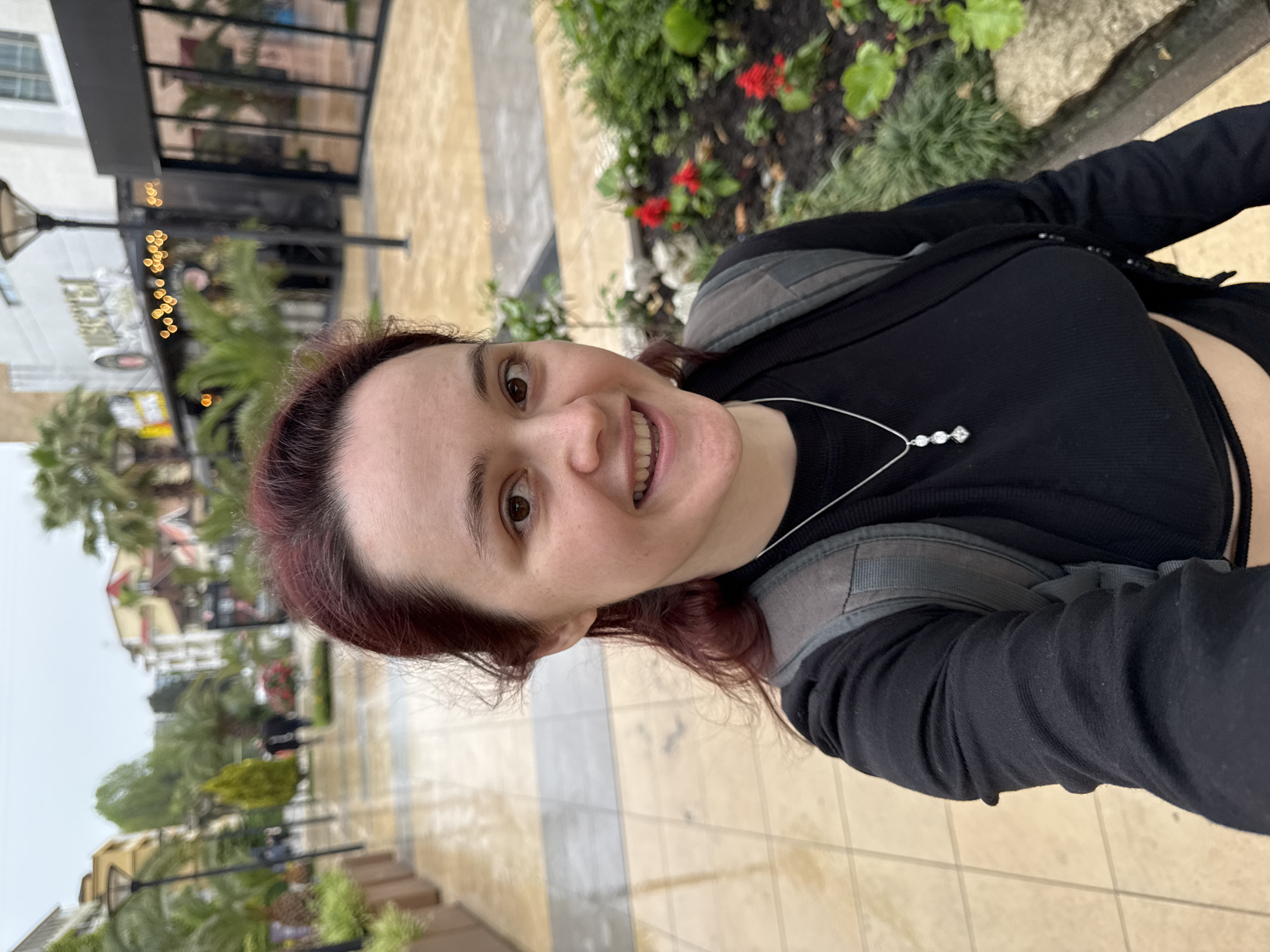 A woman with auburn hair is smiling at the camera, positioned at a slightly upward angle, in Sochi, Russia. She is wearing a black top and a silver necklace with three stones. She also has on a gray backpack. She appears to be standing on a tiled pedestrian walkway lined with plants and flowers, with shops and restaurants visible in the background. The setting suggests an outdoor shopping or dining area. The weather appears to be overcast.