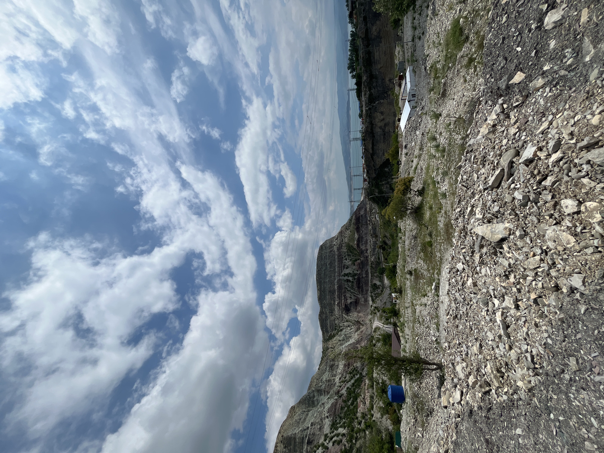 The image shows a landscape with a rocky, mountainous terrain and a cloudy sky, taken near Chirkey, Russia. A slope of rocky material dominates the foreground. In the background, a tall, rocky mountain range is visible under a sky filled with cumulus clouds. A bridge is faintly visible crossing the water in the distance. Power lines stretch across the sky. Vegetation dots the landscape. A blue barrel is near the base of the mountain. In the far background, a settlement or industrial complex is present with long, white buildings.