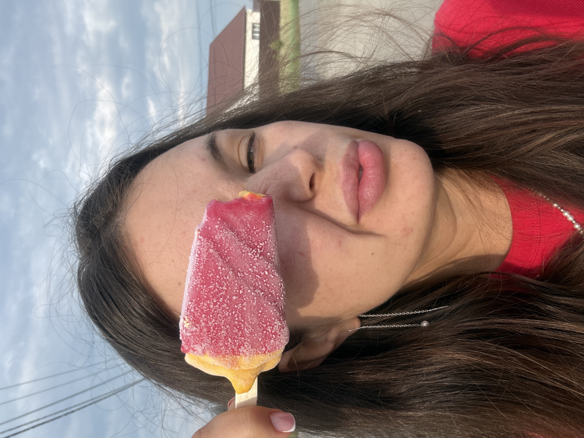 This media file is a portrait of a young woman holding a pink and yellow popsicle in front of her face. She has long, dark brown hair that is falling around her face and shoulders. She is wearing a red top and a silver necklace. Her lips are slightly parted, and her eyes are closed. The popsicle is partially obscuring her face, with the pink part covering her eye and the yellow part visible at the bottom. The background is a cloudy sky with some power lines and a building in the distance, suggesting an outdoor setting. The lighting suggests it is daytime, possibly late afternoon or early evening. The overall mood appears relaxed and casual. There is no visible text in the image.