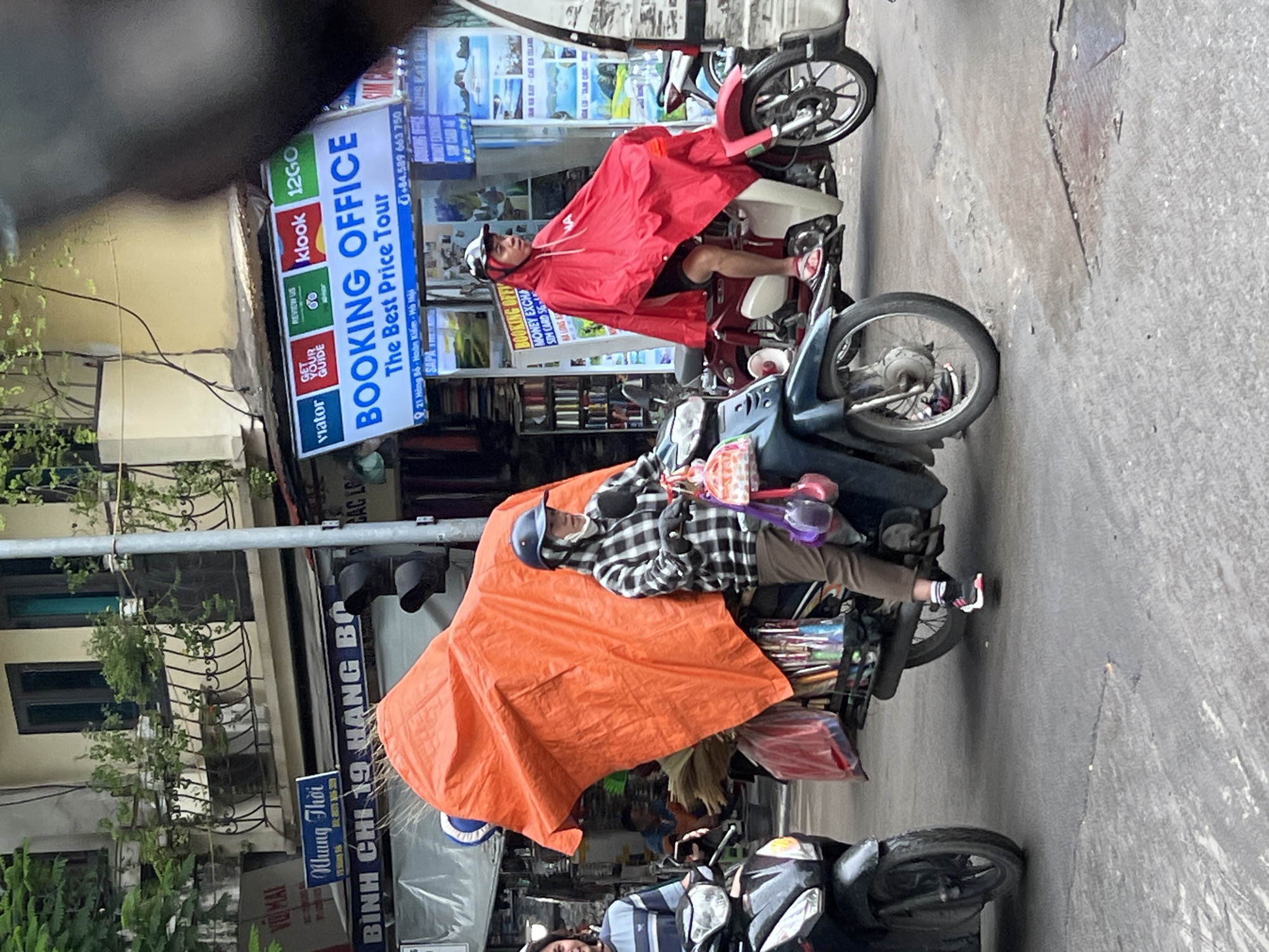 The image depicts a street scene in Hanoi, Vietnam, likely in an urban commercial area. Two people are riding motorbikes. In the foreground, a person wearing a black and white checkered jacket, a blue helmet, and brown pants is riding a motorbike loaded with a large orange tarp covering an assortment of items, including what appear to be brooms and bags. The motorbike has a dark green frame. To the left, another person in a striped shirt is on a black motorbike. Further in the background, a person wearing a red poncho is riding a lighter-colored motorbike. The street is paved with asphalt, and there are buildings and signs visible along the side of the road. One sign reads "BOOKING OFFICE The Best Price Tour." The overall impression is of a busy, everyday scene in a Vietnamese city.