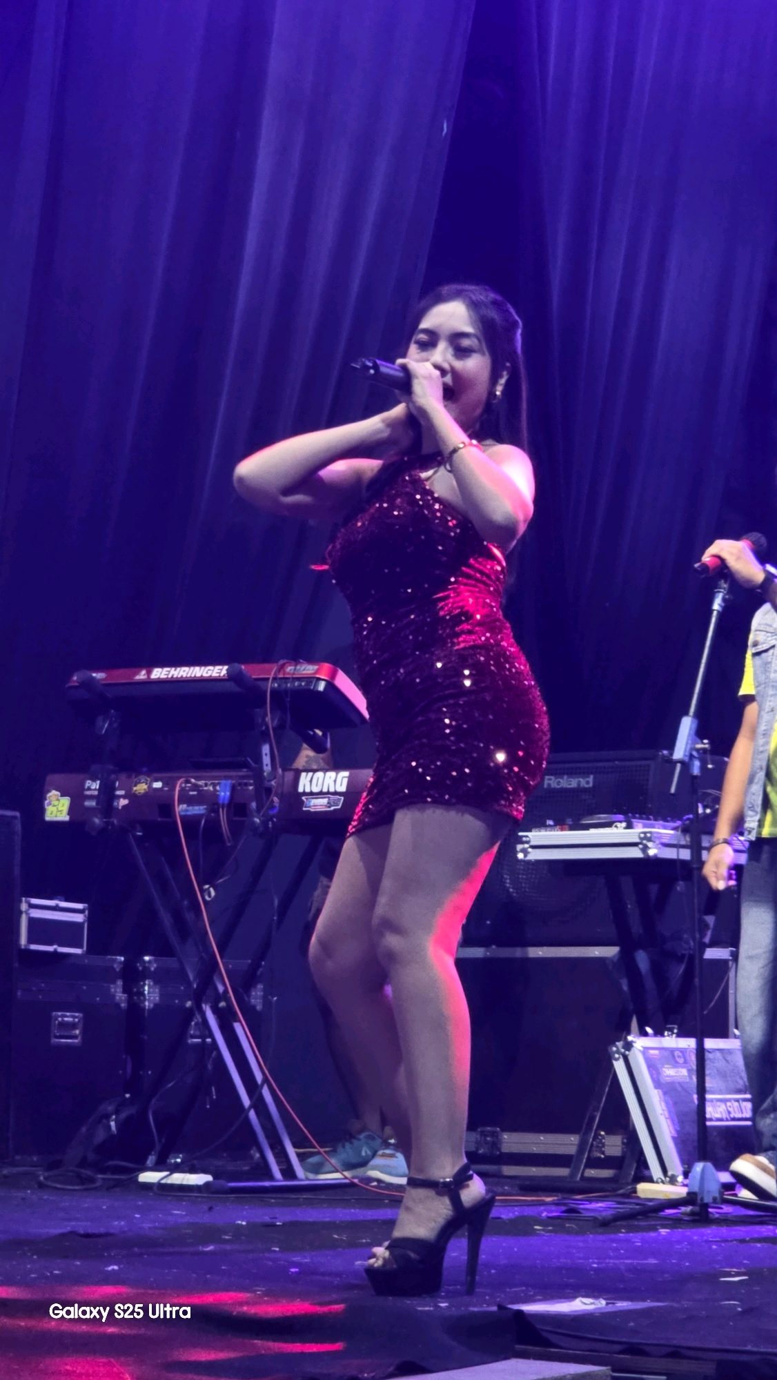 An Indonesian female singer, wearing a shimmering red dress and black high-heeled sandals, performs on a dimly lit stage in Jaken, Indonesia. She is holding a microphone to her lips, her hands near her face. Behind her, a keyboard stand with a KORG keyboard is visible, along with other musical equipment and a dark purple curtain. The stage is illuminated with purple and pink stage lights, casting reflections on the floor. The singer's posture suggests dynamic movement, leaning forward slightly as she sings. The overall scene depicts a live musical performance.