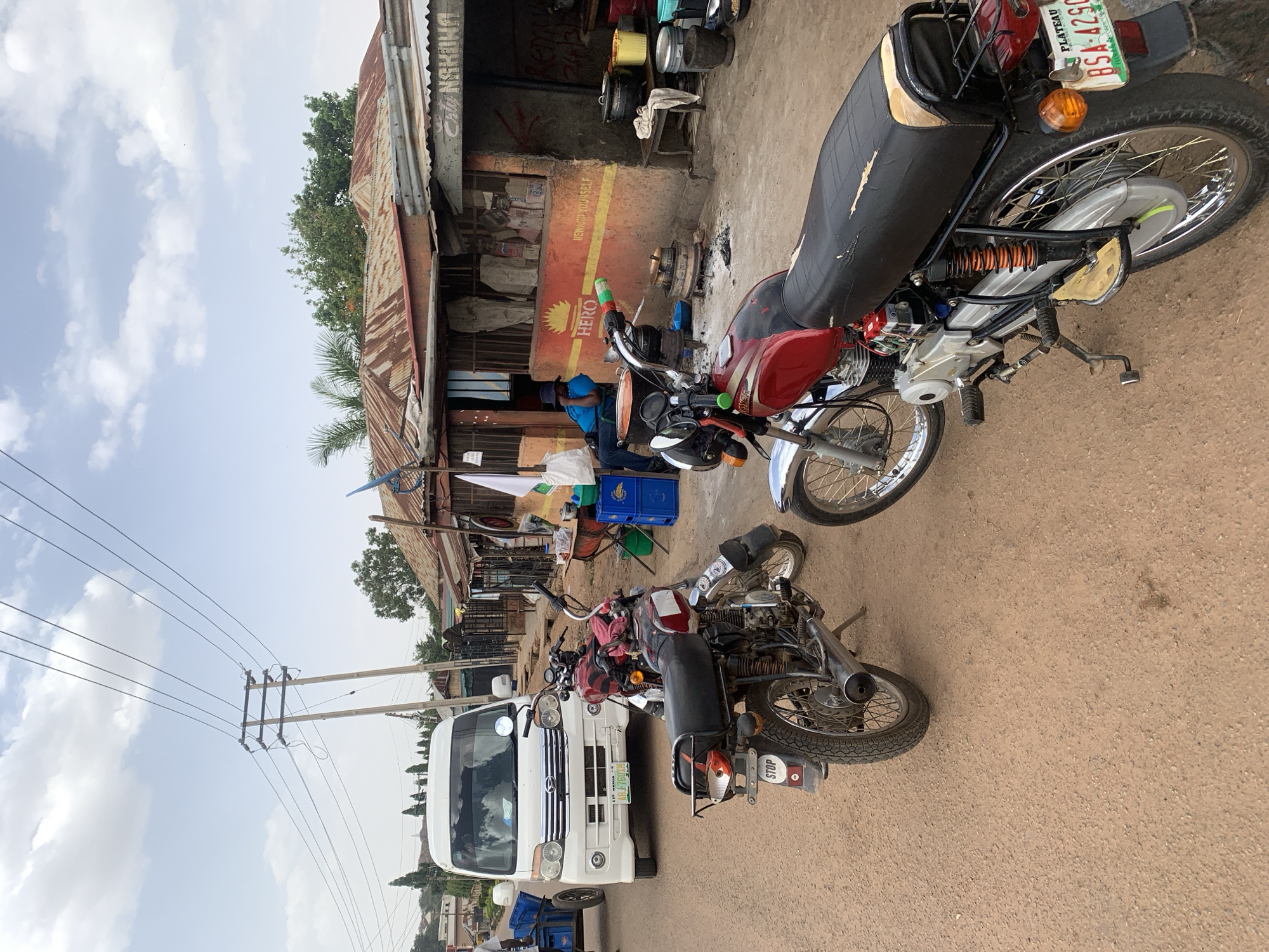 The image depicts a street scene in Jos, Nigeria, featuring several motorcycles parked near a roadside business. A white van is parked on the left side of the road. Utility poles and wires are visible against a cloudy sky. The business appears to be a repair shop, indicated by spare parts and equipment. The scene shows signs of activity.