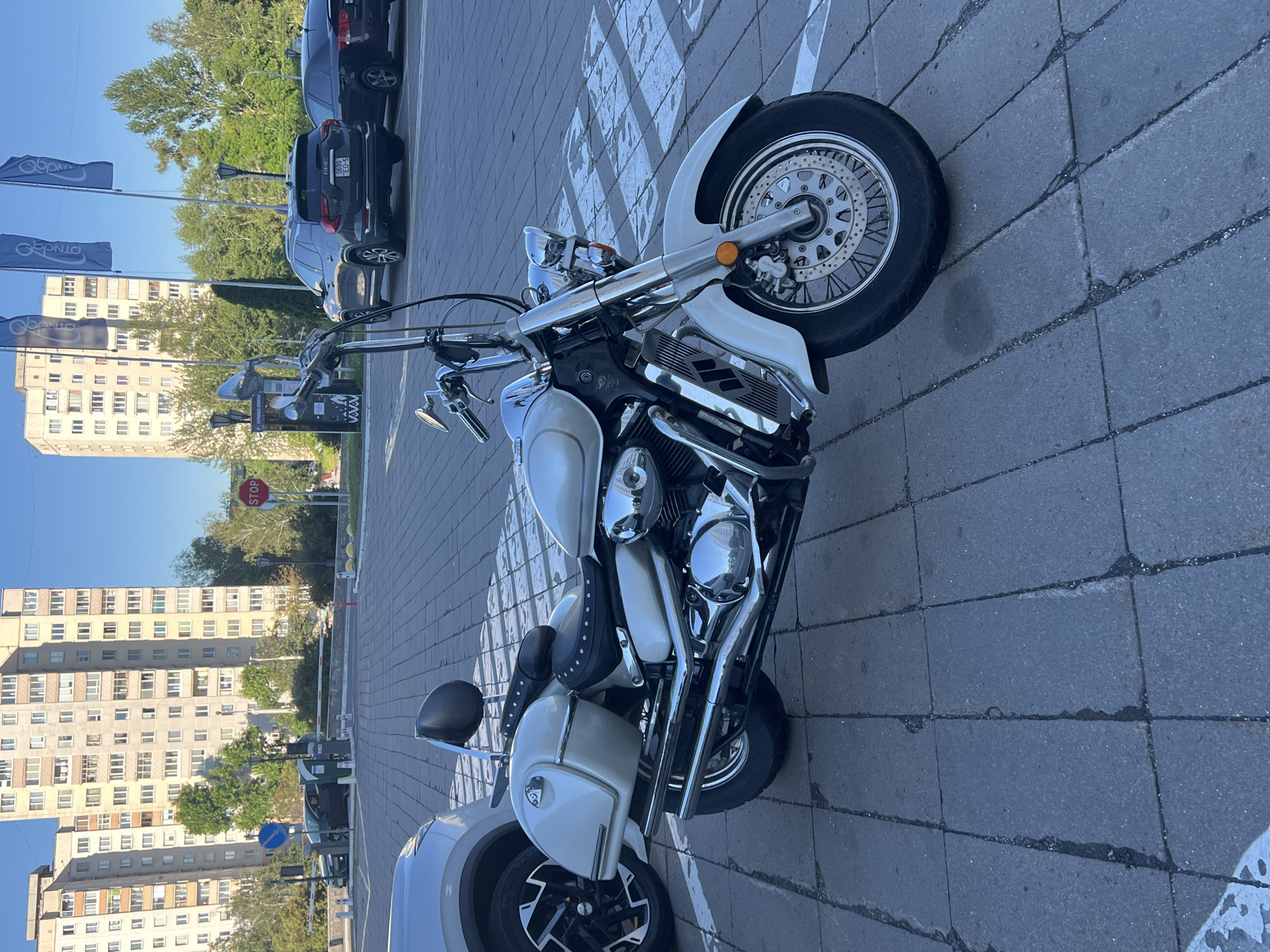 This media file captures a white motorcycle parked on a paved surface, likely a sidewalk or parking lot, in Minsk, Belarus. The motorcycle is a prominent subject, featuring a white body with chrome accents. Its front wheel, handlebars, and engine are clearly visible. The scene includes parked cars in the background, suggesting a public area or residential setting. Tall apartment buildings, characteristic of Minsk architecture, are visible behind the cars, indicating an urban environment. The sky is blue and clear, suggesting daytime and fair weather. A red STOP sign and a blue circular sign with a white arrow are also visible, along with pedestrian crossing markings on the ground. The overall impression is one of a peaceful, everyday scene in a European city. There is no visible text on the motorcycle itself. The flag in the background contains text but it is not legible.