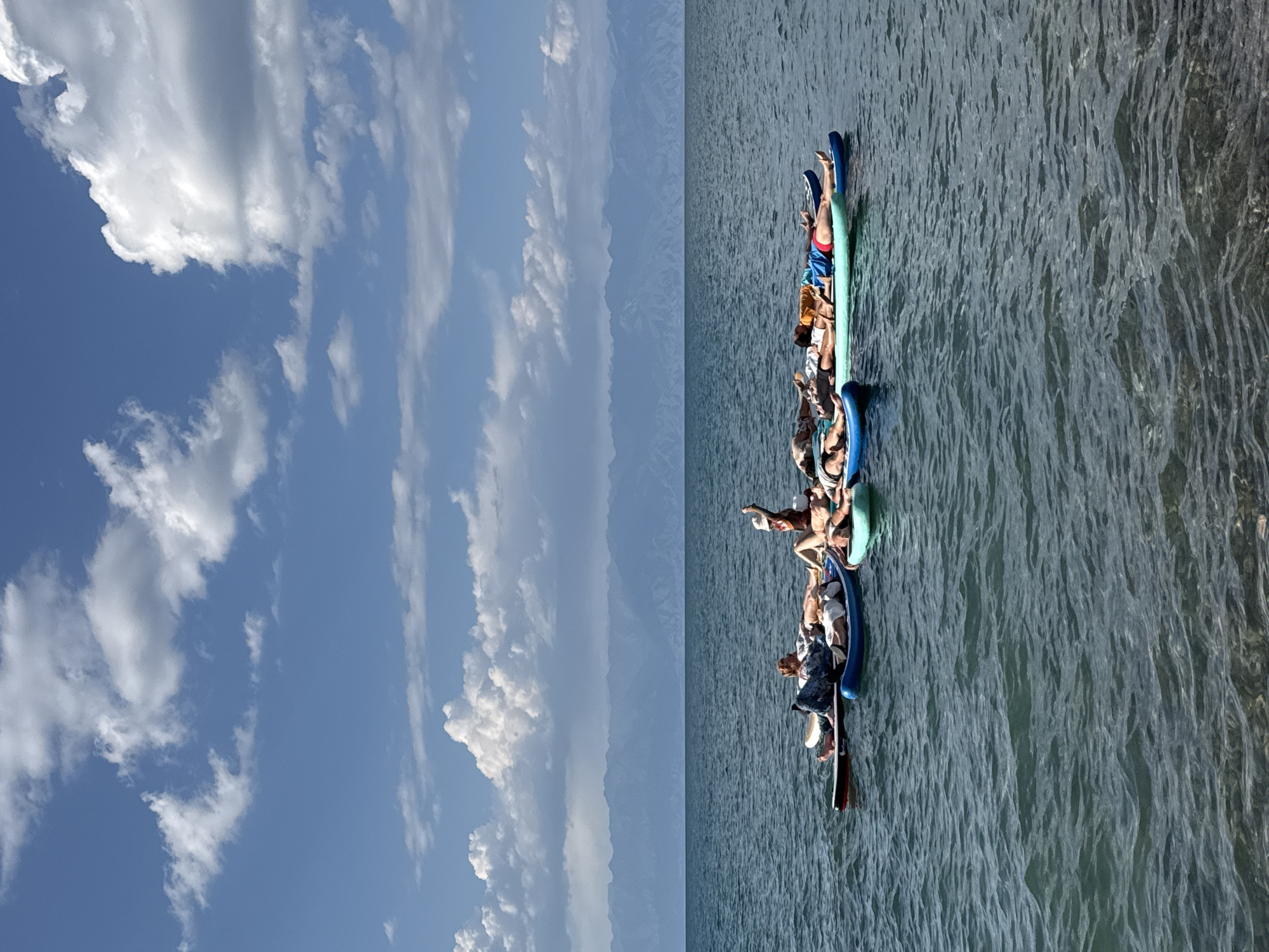 In Basbeshik, Kyrgyzstan, on a clear, sunny day with scattered clouds, a group of people are relaxing on paddleboards in a large body of water. The paddleboards are clustered together, forming a raft-like structure. Most of the individuals are lying down on their backs, some with their eyes closed, seemingly sunbathing or meditating. One person is sitting up on a paddleboard with their arm raised, holding a white object. The water is calm, with gentle ripples, and a distant mountain range is visible on the horizon under a blue sky. The scene appears serene and recreational.
