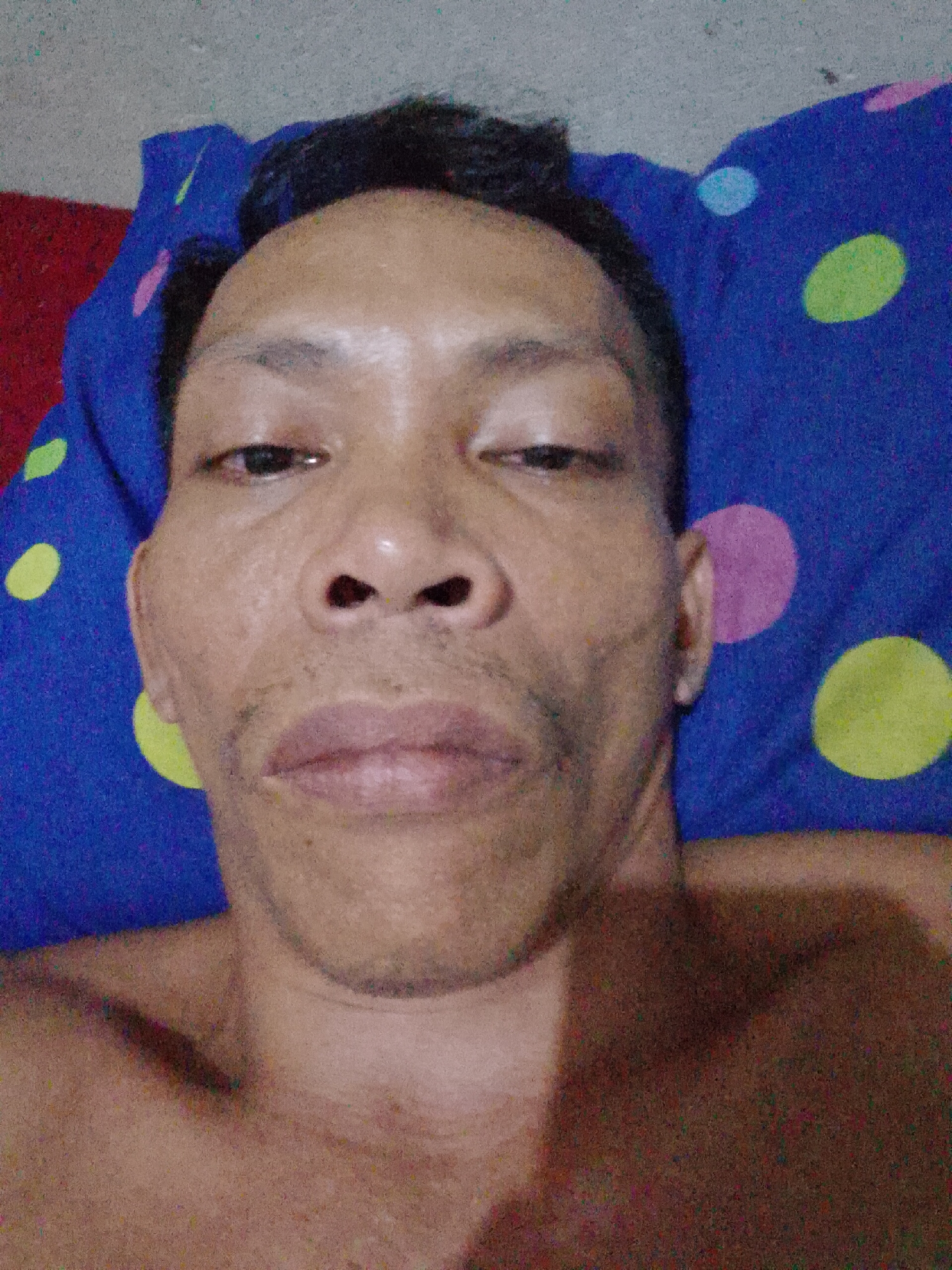 A close-up frontal self-portrait of a man lying on a pillow. The man has dark hair, dark eyes, and a light brown complexion. He appears to be lying on his back. The pillow is blue with colorful polka dots. The background is blurred. The location is Bukit Ketok, Indonesia.