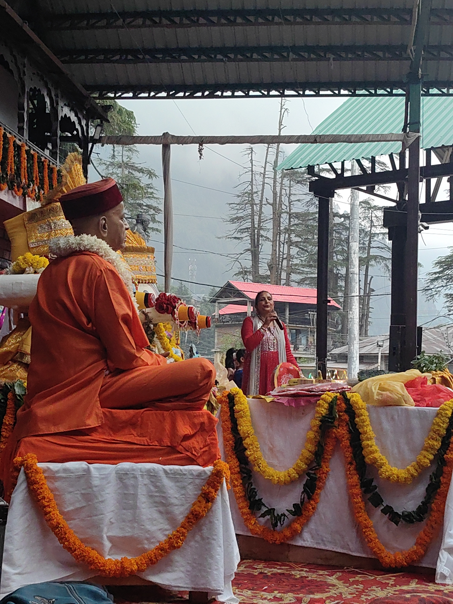 In Manali, India, an outdoor gathering takes place under a covered structure. A man dressed in saffron robes and a maroon cap is seated in the foreground, facing right, with his legs crossed. He wears a garland of white flowers around his neck and has a serene expression. To his right, a woman in a red outfit is speaking into a microphone, facing the seated man and other unseen individuals. She wears a light gray shawl over her shoulders. Behind them, tables are decorated with garlands of orange and yellow marigolds, as well as green foliage. The background reveals tall trees, some bare, against a cloudy sky, suggesting a hilly or mountainous environment. Structures with red roofs are visible among the trees, indicating a village or town setting. The overall scene depicts a religious or cultural event in a natural, elevated landscape.