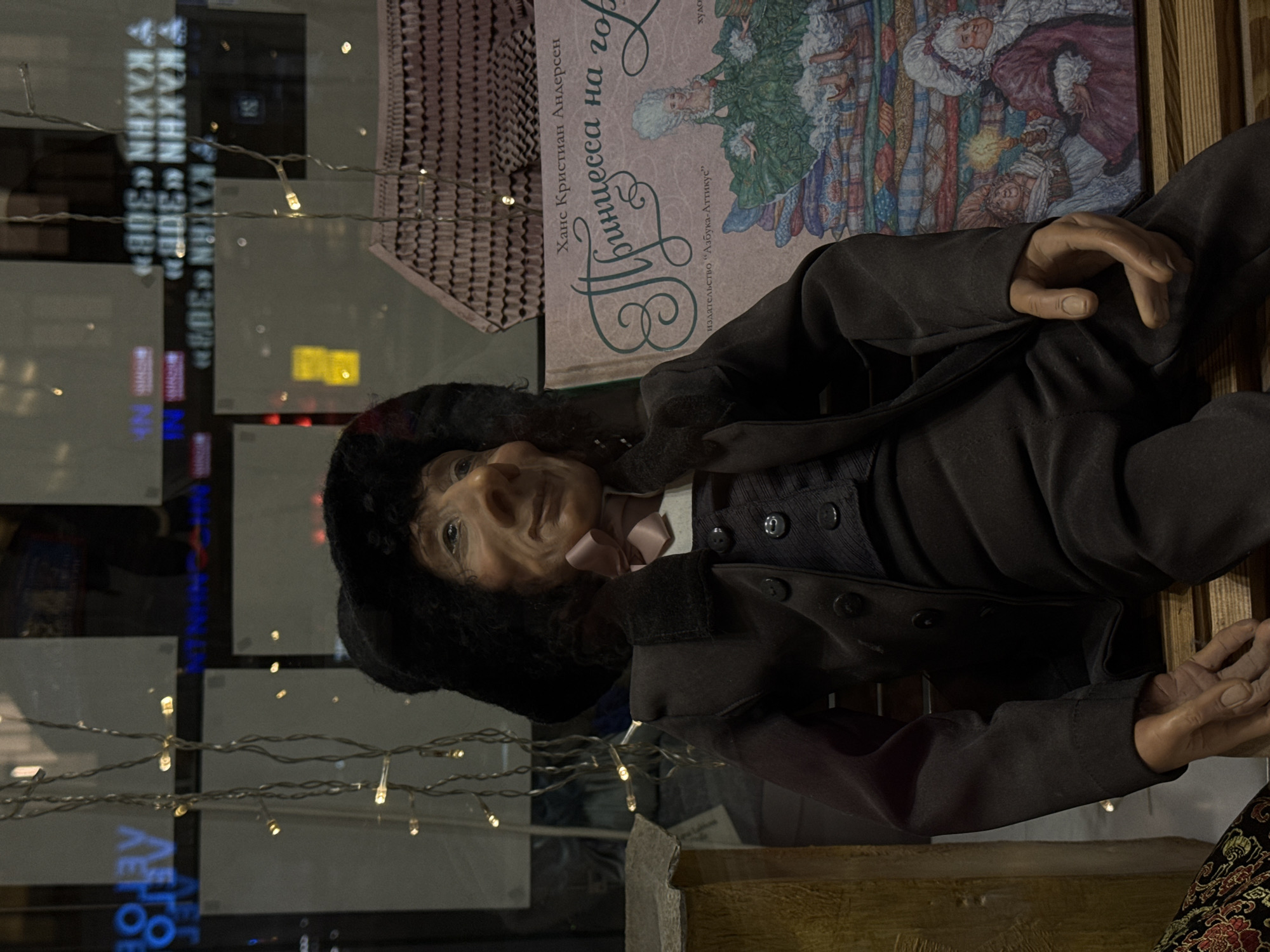 Here's a detailed description of the image:
**Scene Overview:**
The image shows a display, likely in a store window in Moscow, Russia. The display features a mannequin of an older man in historical clothing, posed alongside a book.
**Mannequin Description:**
* The mannequin appears to be a depiction of an older man, dressed in a dark-colored, possibly black, period-style suit with visible buttons.
* He is wearing a fur hat.
* He is wearing a pink bow tie.
* The mannequin's face is detailed with wrinkles and aged features. His hands are realistically sculpted.
**Book Description:**
* Next to the mannequin is a book titled "Принцесса на горо" (Princess on the горо), which is Russian for "Princess and the Pea".
* The book cover displays an illustration typical of fairy tales.
* The name Ханс Кристиан Андерсен (Hans Christian Andersen) is printed on the cover, indicating he is the author.
* The publishing house is listed as "Азбука-Аттикус" ("Azbuka-Atticus").
**Environment:**
* The scene is viewed through a window, as evidenced by the reflections of buildings and lights visible on the glass.
* There are strings of small lights adorning the window, adding to the ambiance.
* Reflections in the window include the text "ΚΑΧΗΝ «308»".
**Location:**
The reflection of the Russian text suggests the photograph was taken in the city of Moscow, Russia.
