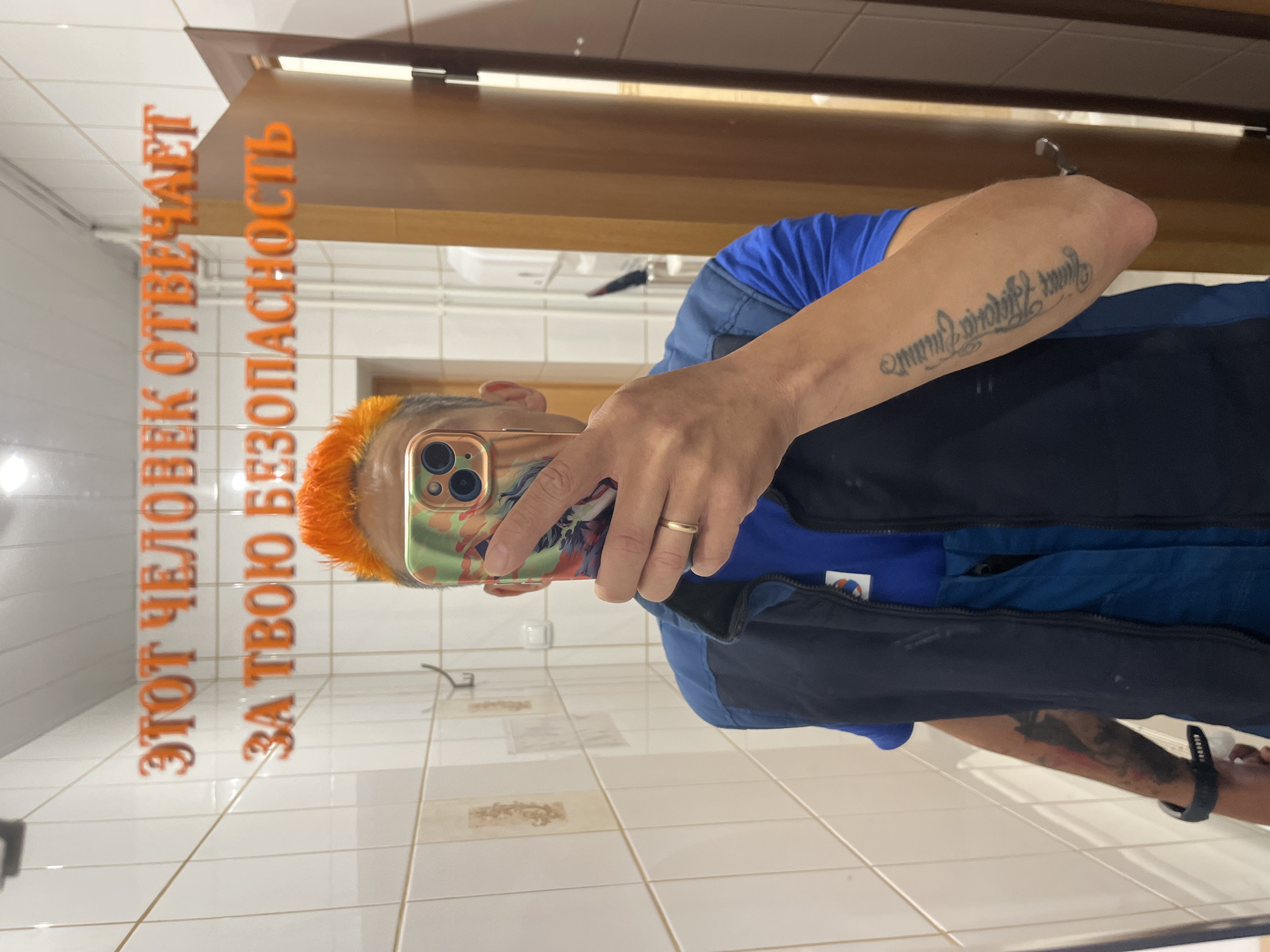 A person is taking a picture of themselves in a mirror. The person has orange hair and is wearing a blue shirt and a dark vest. They are holding a cell phone with a colorful case in their right hand. A gold wedding band is visible on their ring finger. The person has tattoos on both arms. The reflection in the mirror shows a tiled wall and floor, with the Russian text "ЭТОТ ЧЕЛОВЕК ОТВЕЧАЕТ ЗА ТВОЮ БЕЗОПАСНОСТЬ" (THIS PERSON IS RESPONSIBLE FOR YOUR SAFETY) printed on the mirror. The picture was taken in Chaikovsky, Russia.