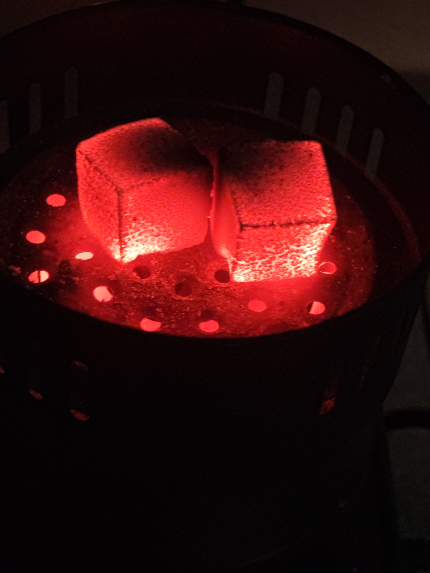 A close-up, overhead shot shows two glowing red charcoal briquettes within a dark metal holder with holes. The briquettes are square-shaped and emit an intense red light that illuminates the surrounding surface, which is also dotted with smaller glowing red holes. The background is predominantly black, with some hints of the perforated rim of the holder visible at the top. The scene appears to be indoors or in a dimly lit environment.
