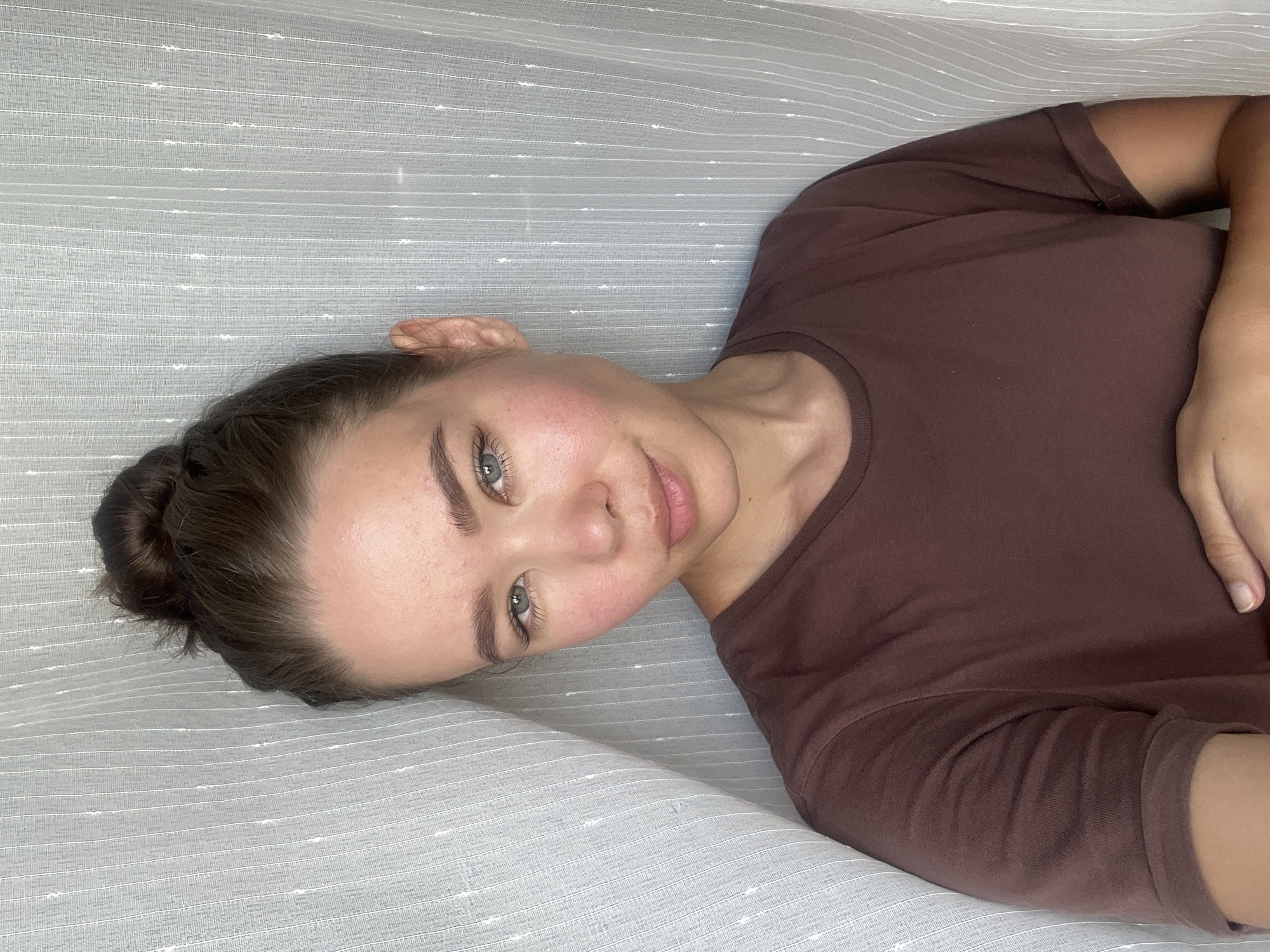A woman with fair skin and light eyes is lying on a surface covered with a light gray fabric with subtle white stripes. Her dark hair is pulled back into a bun. She is wearing a brown t-shirt. The location cannot be determined based on the image provided.