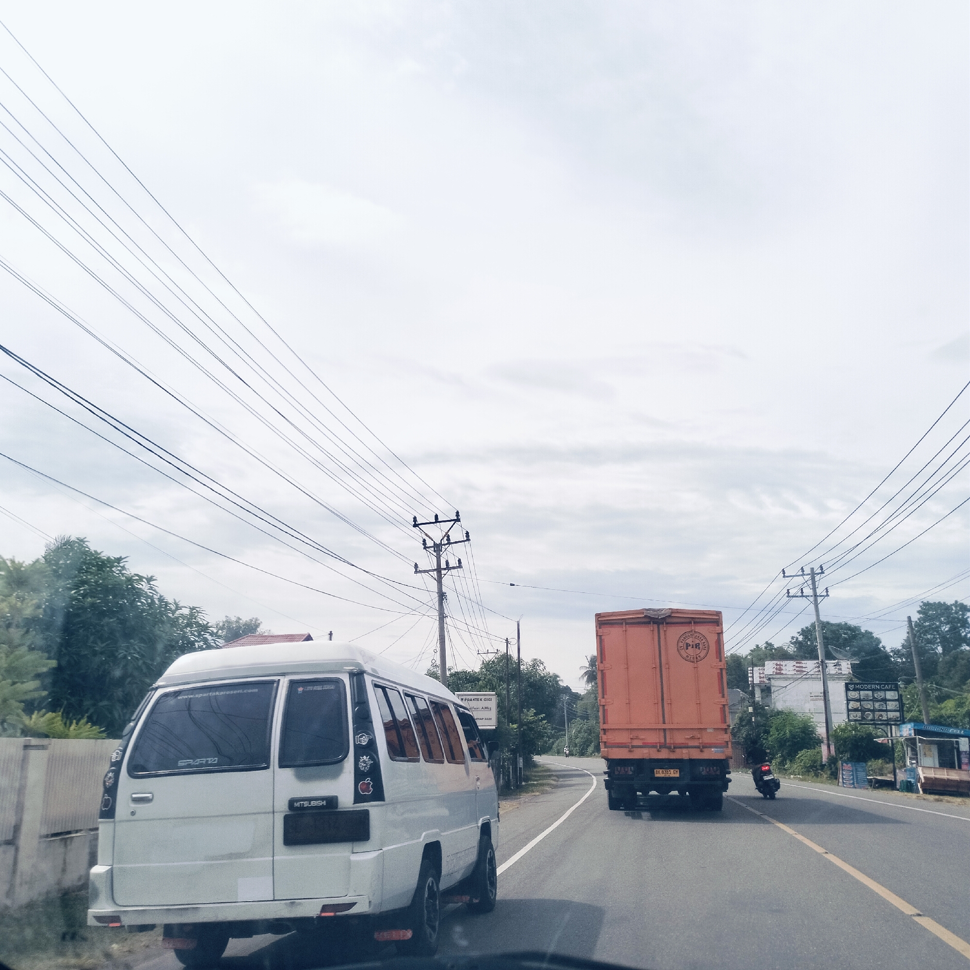 The image captures a road scene from a moving vehicle on an overcast day in Teungoh Baro, Indonesia. In the foreground, a white Mitsubishi van is seen from the rear, featuring "SPARTA" branding and stickers including Apple logos and "www.spartakaroesari.com". Its license plate is obscured. Behind the van, a large orange truck with a "PIA" logo and license plate "BK 8383 GV" proceeds along the road, which has a solid white line and a broken yellow line. A motorcycle trails behind the truck.
Numerous power lines crisscross the cloudy sky, supported by multiple utility poles visible along the roadside. Lush green trees and vegetation line both sides of the road. On the right, a sign for "MODERN CAFE" and a small stall labeled "BURONA CELL" are visible. Further along on the left, a sign reads "PRAKTEK GIGI" (Dentist Practice) with "dr. Afri. AMKg" below. The overall impression is a bustling, semi-urban or rural thoroughfare with everyday traffic and local businesses.