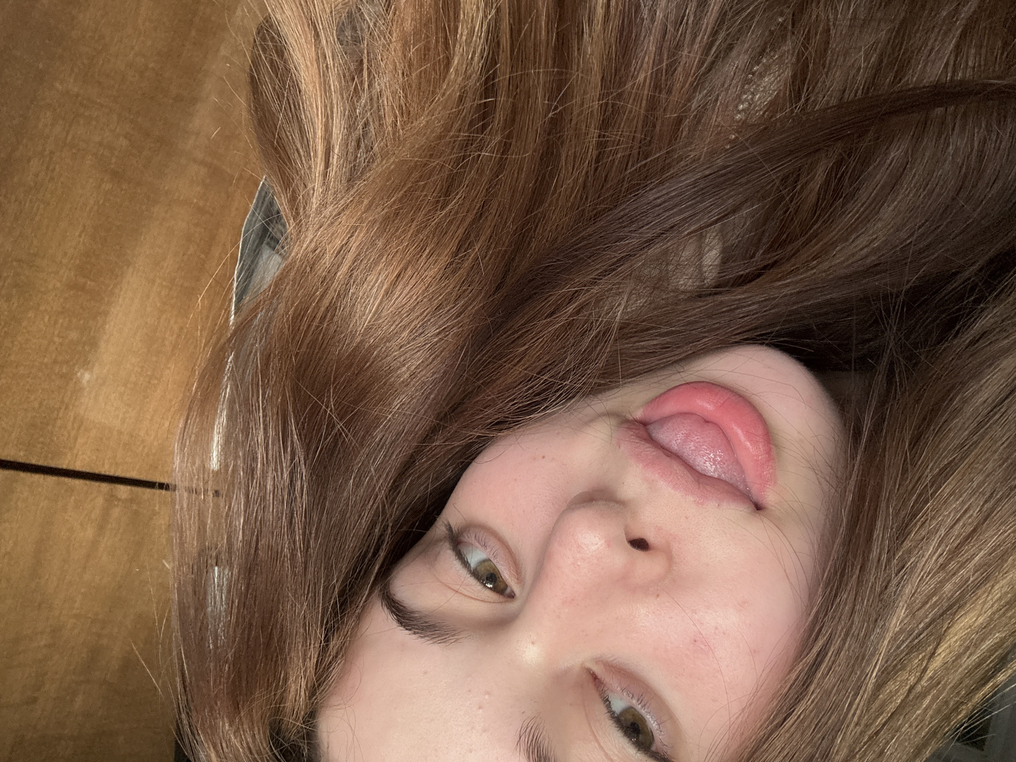 A close-up, angled indoor photograph taken in Troyan, Bulgaria, depicts the upper portion of a young female's face. The subject presents with light brown hair, which frames her face and partially obscures the left side of the frame. Her hazel eyes are directed towards the camera. Her mouth is open, with the tongue extended and slightly curled. The visible skin areas display minor surface variations. The immediate background is a light brown, featureless surface, consistent with a wooden wall or door panel, displaying a faint horizontal line on the right side of the frame.
