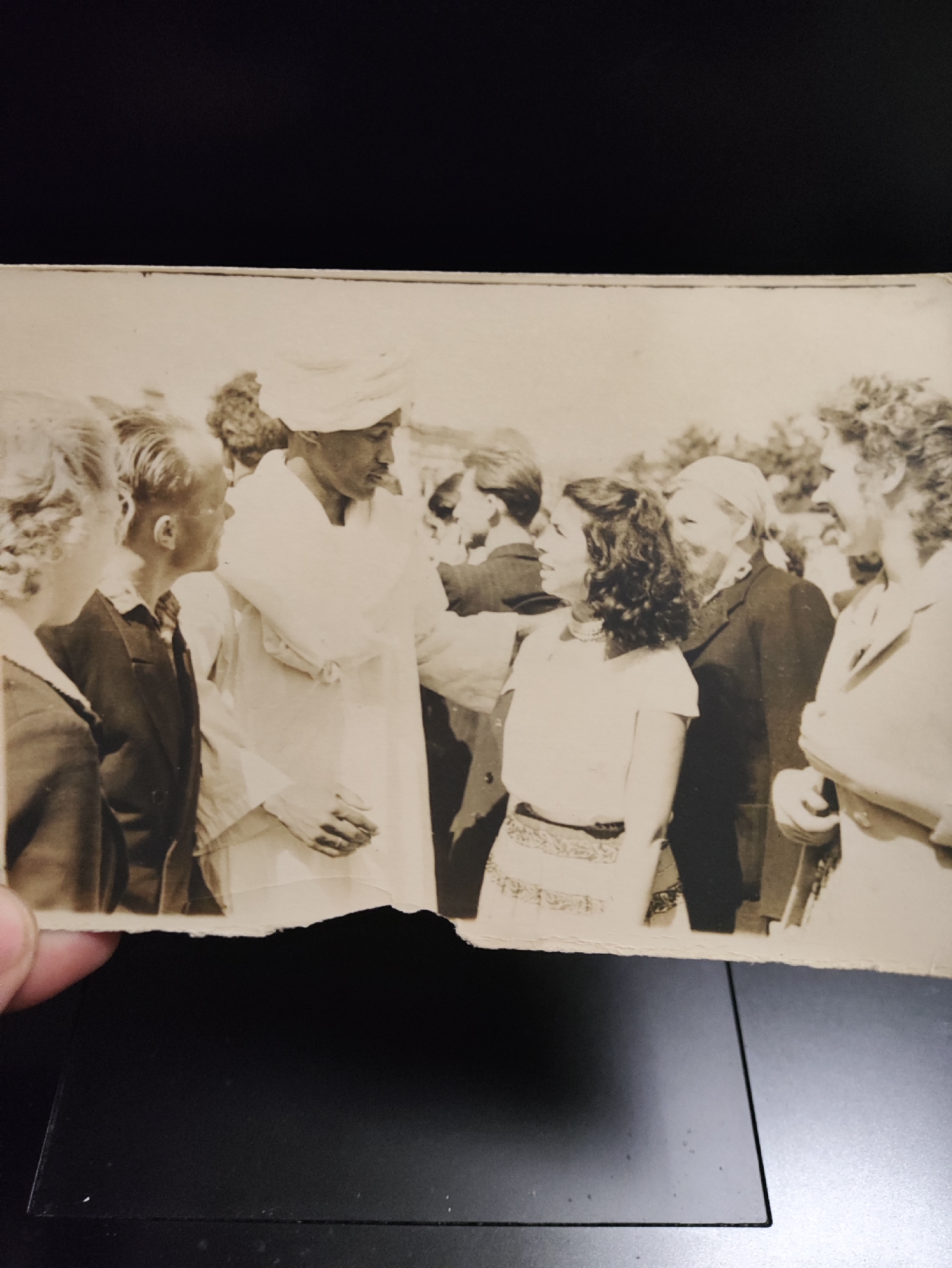 A black and white photograph depicts a group of people standing outdoors, possibly at a public gathering. In the center, a man wearing a white turban and a white robe is engaged in conversation with a woman who has curly dark hair and is wearing a light-colored dress. To their left, two individuals are partially visible. One is a young boy in a dark suit, facing the man in the turban. To his left, a woman with curly hair is also partially visible. To the right of the central figures, an older woman with light-colored hair and a dark jacket is looking towards the central woman. Further to the right, a woman with curly hair and a light-colored jacket is smiling and looking towards the left. The background consists of blurred foliage and other indistinct figures, suggesting an outdoor setting. The photograph is torn along the bottom edge. There is no information in the image to suggest it is located in Izhevsk, Russia.