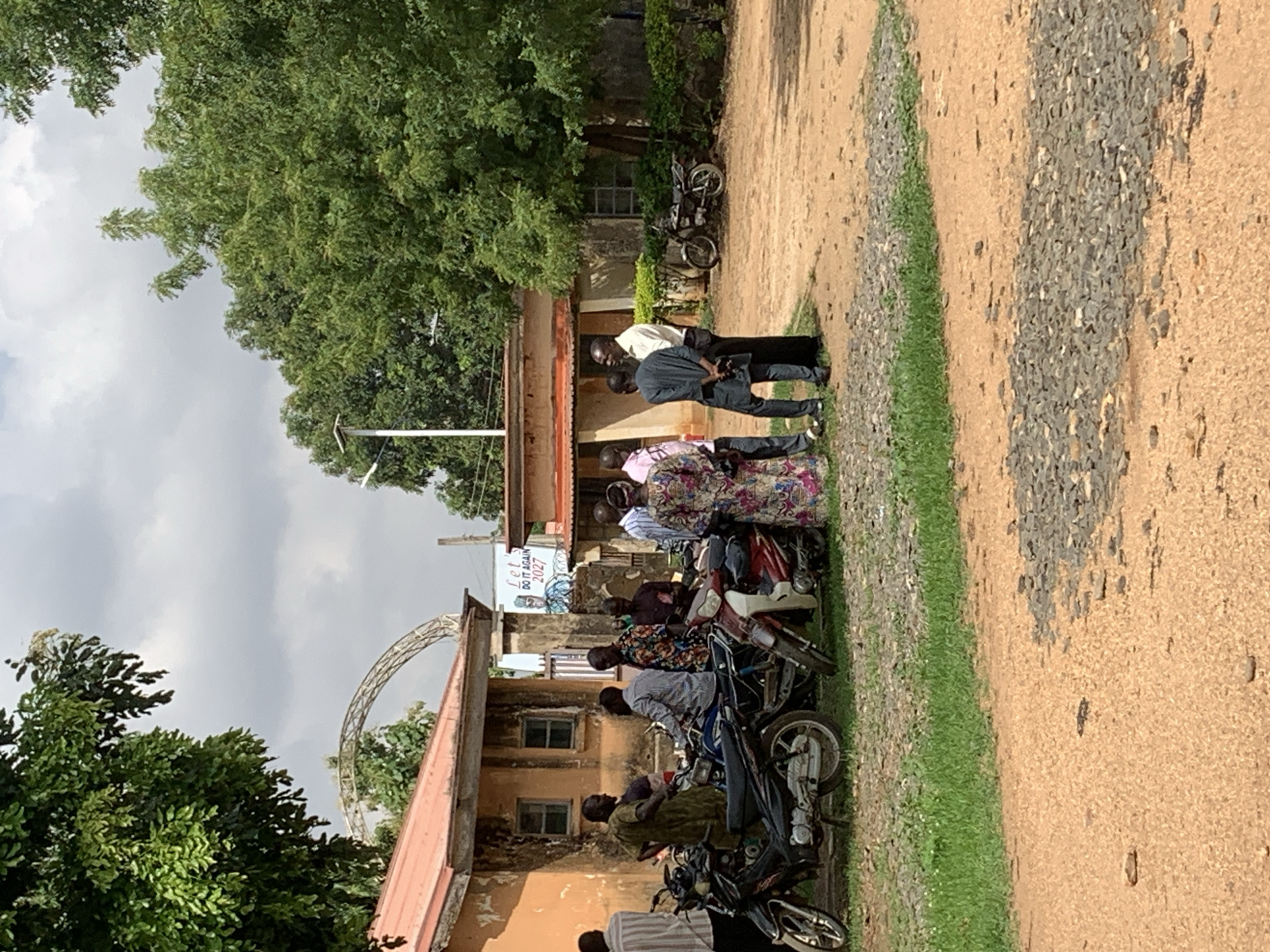 In Maigizo, Nigeria, a group of approximately 10 individuals is gathered near a building. Several motorcycles are parked along the side of the building. A sign with the words "Let's Do it Again 2027" is visible. Trees and foliage surround the building. A curved metal structure stands atop the building's roof. The ground is dirt and gravel, with a strip of grass. The sky is overcast.