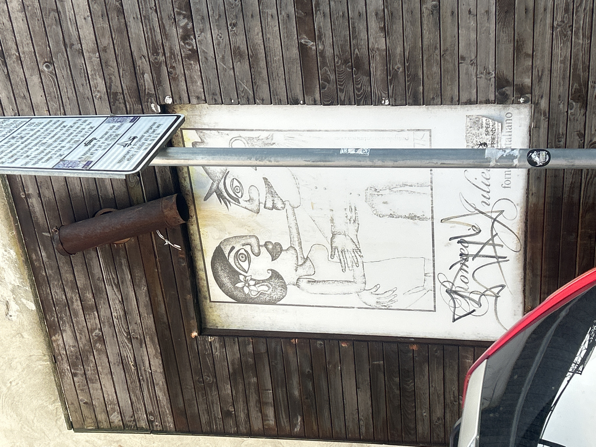 The image shows an outdoor scene in Burgas, Bulgaria. A weathered wooden structure serves as a backdrop. On it hangs a black-and-white drawing depicting two figures with distorted features. Below the drawing, the word "Romeo" is visible, accompanied by additional script that includes the phrase "from Italiano." A metal pole cuts through the image horizontally, supporting a sign with text and graphics. A rusty pipe is attached to the structure. The rear side of a car is visible on the right.
