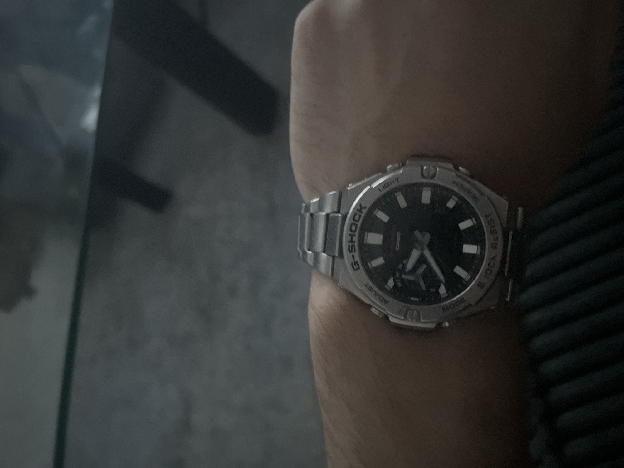 The image shows a close-up of a person's wrist wearing a silver G-Shock watch with a black face. The watch has luminous hour markers and hands, and the brand name "G-SHOCK" is clearly visible on the bezel. Other text on the watch includes "LIGHT", "ADJUST", "MODE", and "START/STOP". The watch band is also silver and appears to be made of metal. The background is out of focus, but it appears to be an indoor setting with a dark, textured surface and some darker, blurred shapes that could be furniture. The lighting in the image is dim, suggesting it might be nighttime or an overcast day. The person's skin is visible on the wrist, and it appears to be a light skin tone with some light hair. The image is cropped tightly on the wrist and watch, with little of the surrounding environment visible.