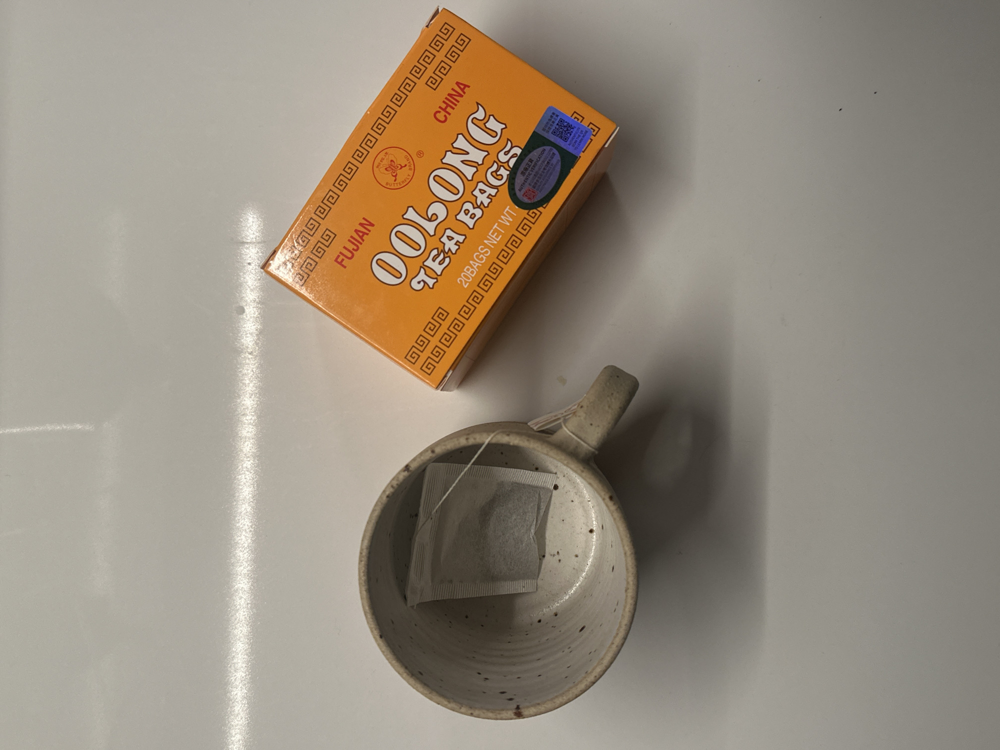 The image shows a tea box of Fujian Oolong tea bags and a mug with a tea bag inside on a white surface. The tea box is orange with black lettering and a holographic sticker. The mug is white with brown speckles and a visible tea bag inside. The setting appears to be indoors.