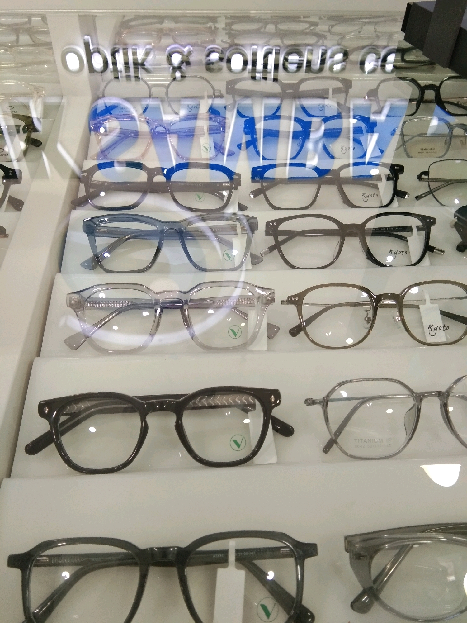 A display case filled with various eyeglasses is shown. The eyeglasses are arranged on multiple shelves and are of different styles and colors, including black, blue, and clear frames. Some of the eyeglasses have brand names visible on them, such as "Kyoto" and "TITANIUM IP". The display case appears to be in a retail setting, possibly an optical shop. The text "obrik & eoueo cc" is reflected on the glass surface of the display case. The image quality is clear, and the lighting is bright, illuminating the eyeglasses.