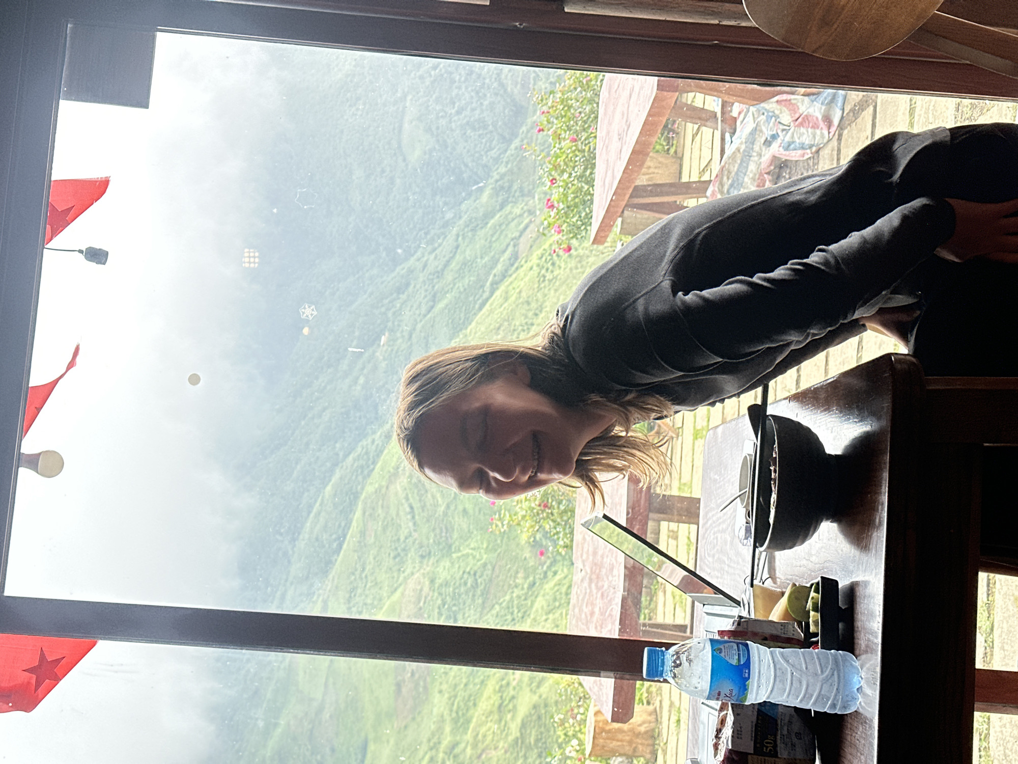In Ta Xua, Vietnam, a young woman with braces and light brown hair is captured in a moment of laughter, leaning forward. She is wearing a dark grey long-sleeved shirt. In front of her on a dark wooden table, a bottle of water, a black bowl of food, and a small plate with sliced lime are visible. The table is positioned by a large window that offers a panoramic view of lush green mountains shrouded in mist. Red flags with stars are attached to the top frame of the window, and outdoor seating areas with wooden tables and benches are visible through the glass.