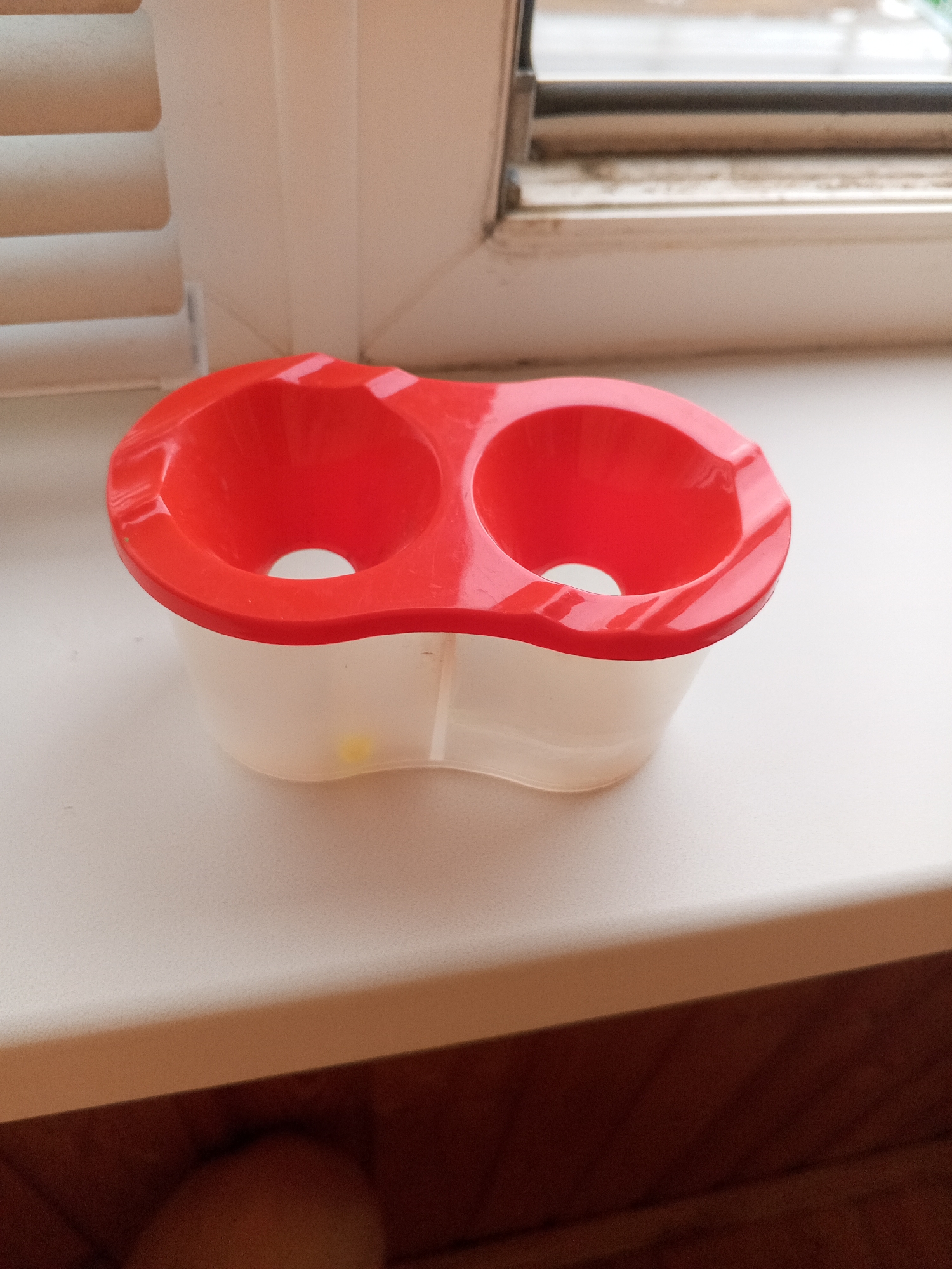 A red and translucent plastic egg holder with two indentations sits on a white windowsill. To the left of the egg holder are closed white blinds. Behind the egg holder and blinds is a window with a dirty sill. The windowsill appears to be made of wood. The image was taken in Chistopol, Russia.