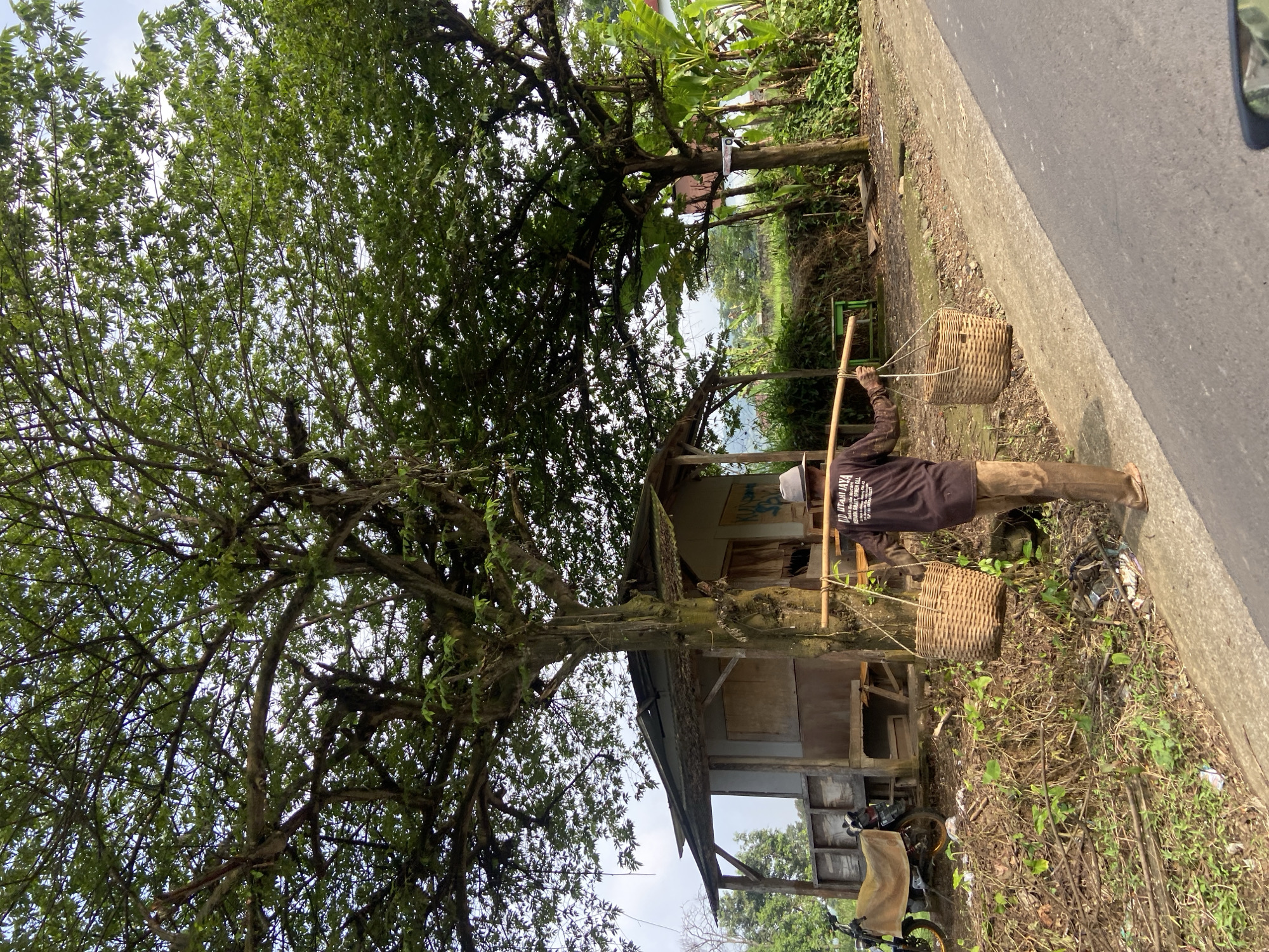 A man carries two baskets suspended from a pole across his shoulders on a rural roadside in Sukoreno, Indonesia. He is dressed in a dark long-sleeved shirt and light-colored pants. A small wooden structure stands nearby, partially obscured by a large tree with dense foliage. A bicycle is visible next to the structure. A paved road runs along the edge of the scene. The background includes vegetation and a glimpse of a distant landscape.