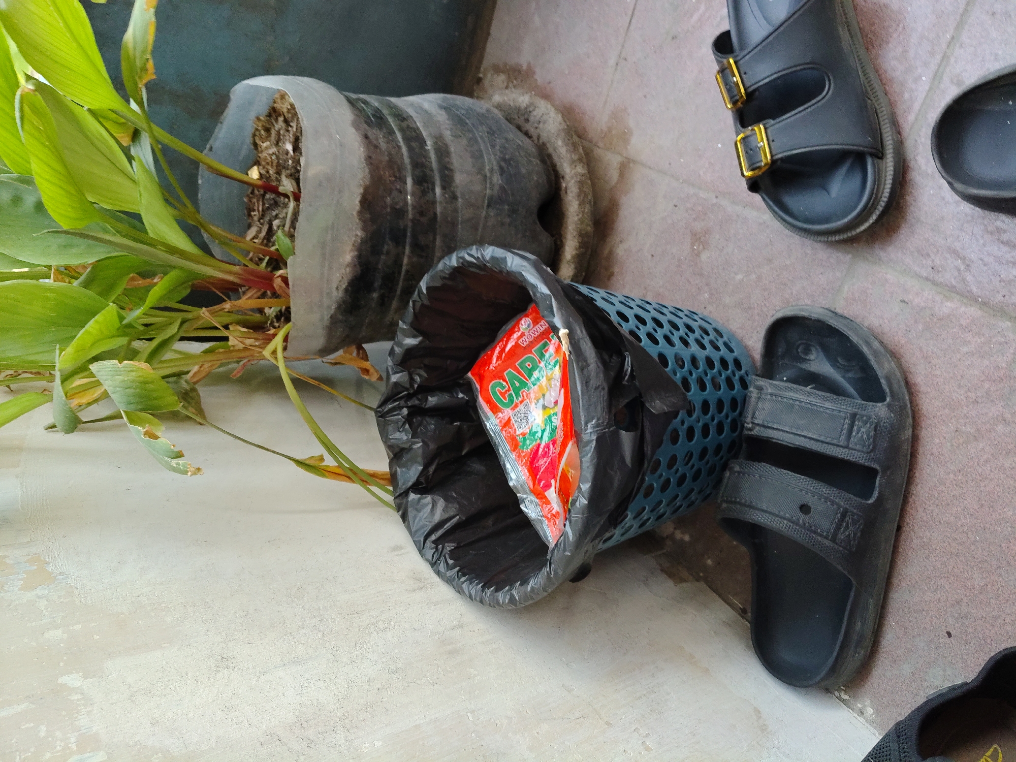 The image captures an outdoor scene with a light-colored, distressed wall on the left and a potted plant in a makeshift container on the right. In front of the wall is a blue plastic basket with a black trash bag inside. A bright orange and red packet is visible within the trash bag. In the foreground, there are three pairs of black sandals on a tiled floor. The scene is situated in a typical residential area in Surabaya, Indonesia, likely a backyard or patio. The lighting suggests it is daytime, possibly with diffused sunlight.