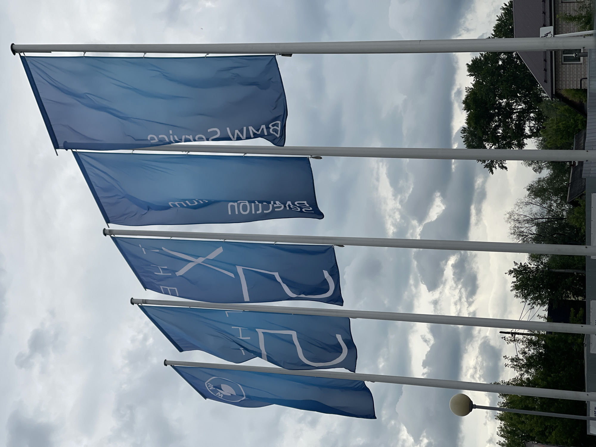 The image captures a set of five blue flags flying vertically from white flagpoles against a cloudy sky. The photograph is taken from a low angle, looking upwards. The flags are gently waving, suggesting a light breeze.
Reading the text on the flags from top to bottom (as displayed in the image, noting that the text appears mirrored on the fabric), the following phrases are visible:
1. "BMW Service"
2. "BMW Premium"
3. "THE X" (followed by partially visible characters, likely indicating a BMW X-series model)
4. "THE M" (followed by partially visible characters, likely indicating a BMW M-series model)
5. The circular BMW logo.
In the background, behind the flagpoles, there are green trees and a building with a dark-tiled roof and light-colored walls. A sign featuring a white wheelchair accessibility symbol on a purple background is visible near the building. The sky is overcast with white and gray clouds, indicating daytime. A white street lamp with a spherical light fixture is also visible in the lower right portion of the background. The scene suggests an outdoor commercial setting, likely a BMW dealership or service center.