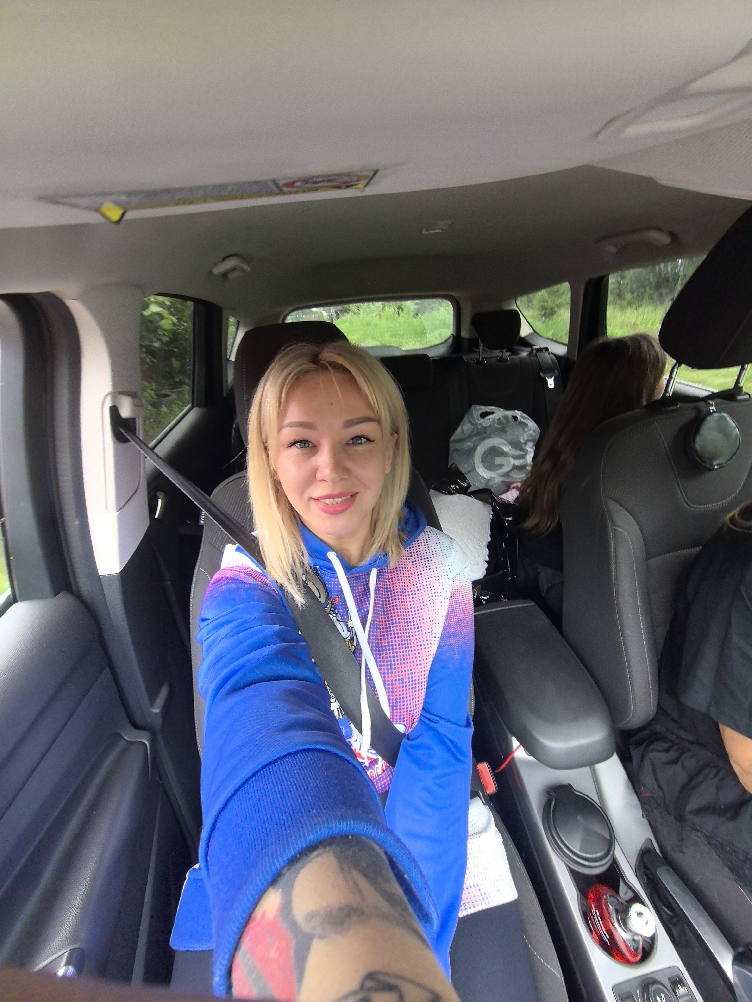 The image shows a blonde woman with blue eyes and pink lipstick sitting in the passenger seat of a car, taking a selfie. She is wearing a blue hoodie with pink and white accents and a seatbelt. Her left arm, which is visible in the foreground, is tattooed. In the background, two other individuals are seated in the back of the car. The interior of the car appears to be gray and black. Outside the car windows, lush green trees and foliage are visible, suggesting a natural or rural setting. The scene likely takes place on a road in or near Noviy, Russia, given the context provided.