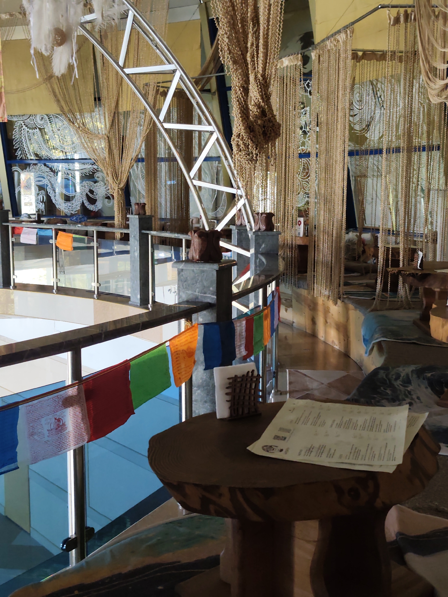 The image depicts the interior of a cafe or restaurant in Sochi, Russia, characterized by its unique design and decorative elements. A prominent feature is the extensive use of hanging beads and strings, creating a curtain-like effect throughout the space. These hangings are a light golden-brown color and vary in thickness and density, contributing to a warm and somewhat enclosed atmosphere.
In the foreground, a round, wooden table with a textured surface, resembling a slice of a tree trunk, is visible. On this table, there are two pieces of paper, likely a menu, with printed text and some logos. To the left of the table, a railing is adorned with a string of prayer flags in various colors, including blue, white, red, green, and orange. The flags are small and appear to be made of fabric.
Further into the scene, a modern, curved metal structure, possibly part of the building's architecture or a decorative element, arches over the space. Behind this structure, large windows with intricate white patterns are visible, suggesting artistic decoration on the glass. More hanging bead curtains are present, framing the windows and the general area.
The environment appears to be elevated, with a glimpse of a lower level through the railing, showing blue glass walls. The overall ambiance suggests a blend of modern design with traditional or spiritual decorative touches. There are no people clearly visible in the image. The lighting is a mix of ambient light and possibly some artificial sources, creating soft shadows.