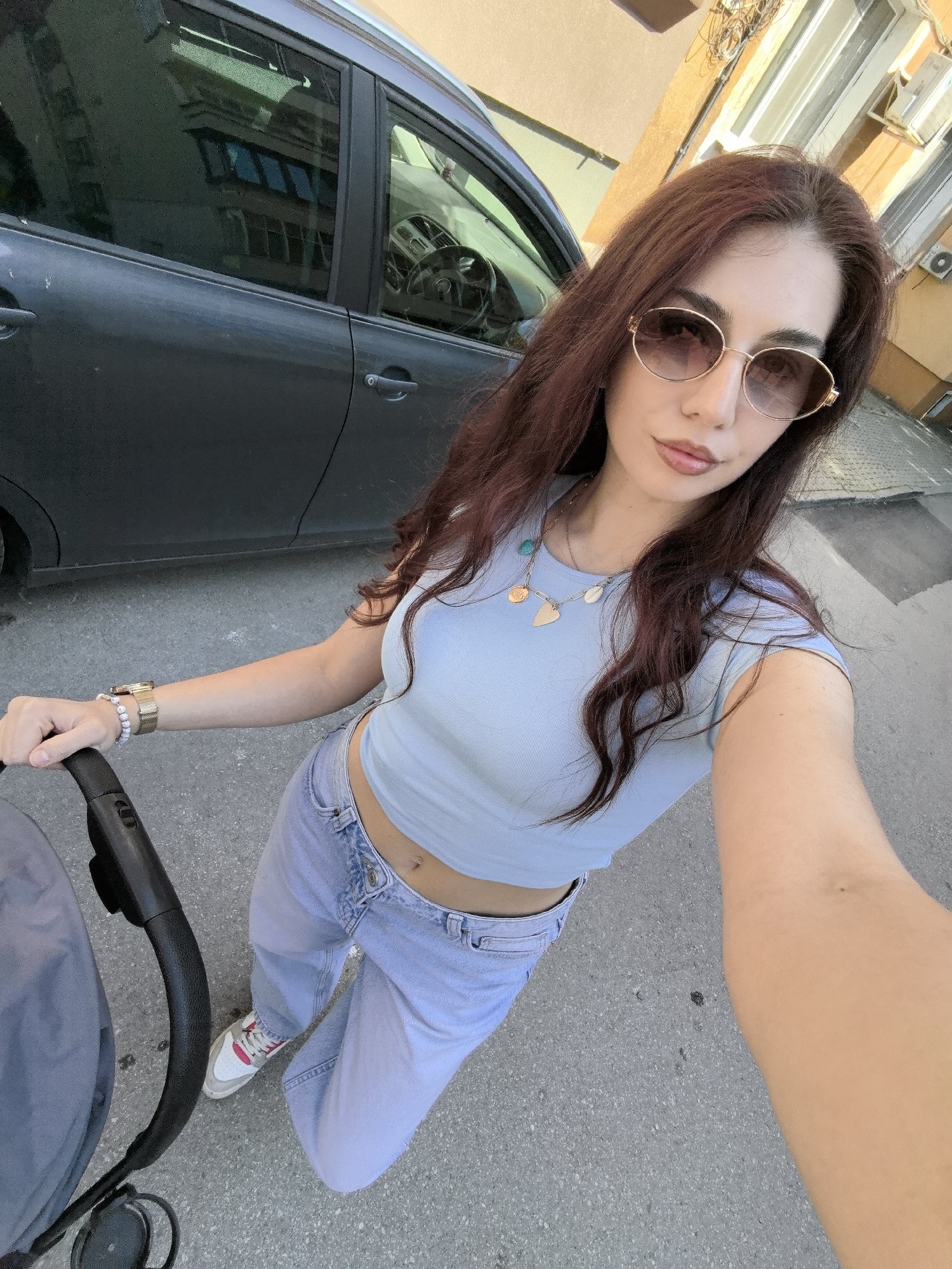 A woman with long, auburn hair, wearing oval sunglasses, a light blue crop top, and light blue jeans, is holding a baby stroller. She is standing on a gray asphalt road. Behind her is a dark gray car parked beside a building. The building has a yellow facade and white window frames. The location is likely in Stara Zagora, Bulgaria.