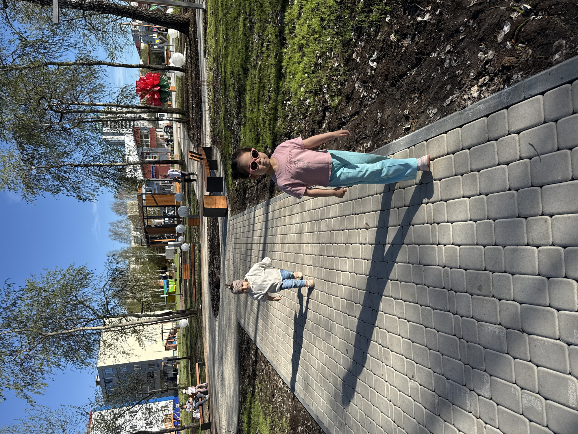 A photograph, taken in a park in Tobolsk, Russia, shows two children walking on a paved walkway. One child, wearing a pink t-shirt, blue pants, and pink sunglasses, is in the foreground. The other child, wearing a light-colored jacket, blue jeans, and a beige head wrap, is slightly behind the first child. The walkway is made of gray paving stones. A grassy area runs alongside the walkway, bordered by bare dirt. In the background are trees, buildings, and park benches with people sitting on them. The sky is blue.