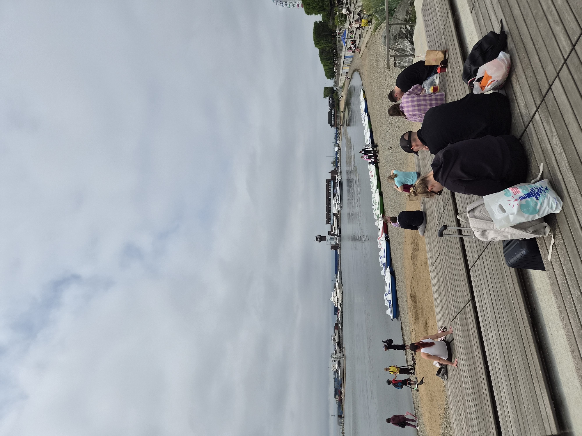 The image depicts a view of the waterfront in Vladivostok, Russia, on a cloudy day. The foreground is dominated by a wooden deck with several people seated on it, facing away from the camera. Luggage, including suitcases and bags, is visible on the deck.
Beyond the deck is a pebble beach where more people are gathered. On the beach, there are several pedal boats lined up near the water's edge. The water is calm, and in the middle ground, a harbor is visible with various boats and yachts docked. A distinctive tower, possibly a lighthouse or control tower, stands near the harbor.
The background shows more of the city's skyline with buildings and trees, and a Ferris wheel is visible in the distance to the right. The overall atmosphere suggests a relaxed day by the sea, with people enjoying the waterfront.