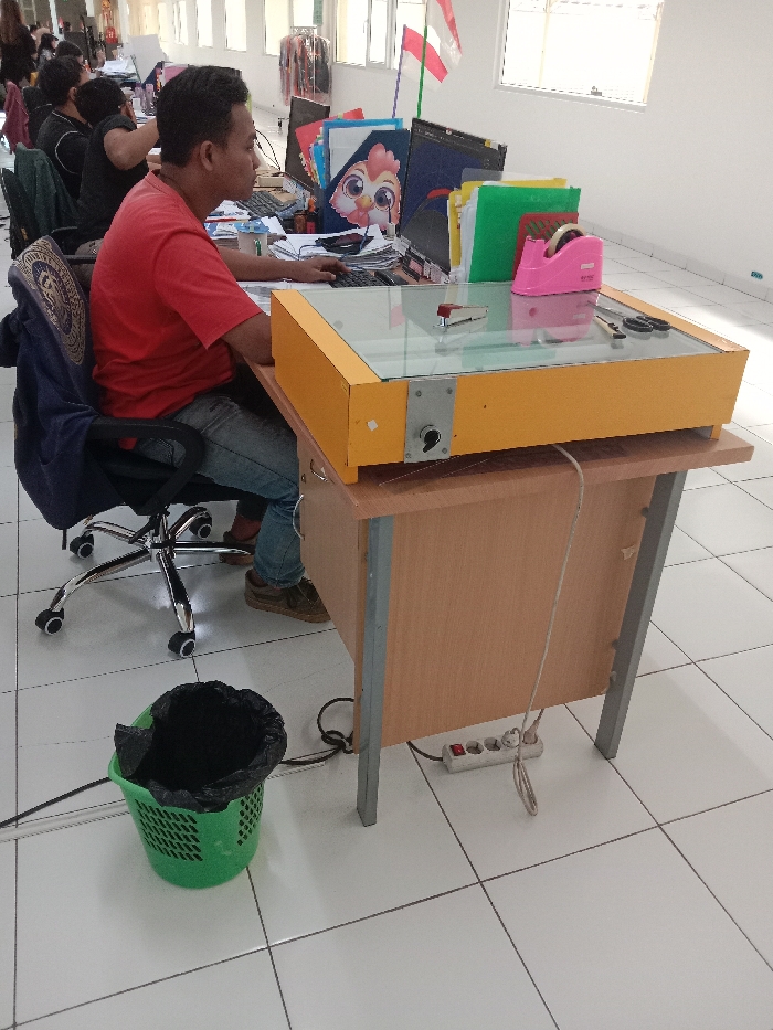 The image shows a man sitting at a desk in an office. The man is wearing a red shirt and blue jeans, and he is using a computer with a yellow-and-glass top. He is positioned in a cubicle, which is lined with white tiles. The cubicle is in a larger office, which appears to have large windows with a view of another building. There is a black trash can with a green liner to the man's left. The image was likely taken at an office in the Kecamatan Ngaliyan district of Semarang, Indonesia, based on the provided coordinates. A power strip is visible on the floor underneath the desk. Several colorful binders are visible on a shelf behind the man. A red-and-pink tape dispenser sits atop a stack of orange and green binders. A red object is visible on the surface of the desk, in front of the man. The image is taken from a slightly elevated angle, and the focus is on the man and his desk. The image appears to have been taken in natural light. There is no visible interaction between the man and any other people.