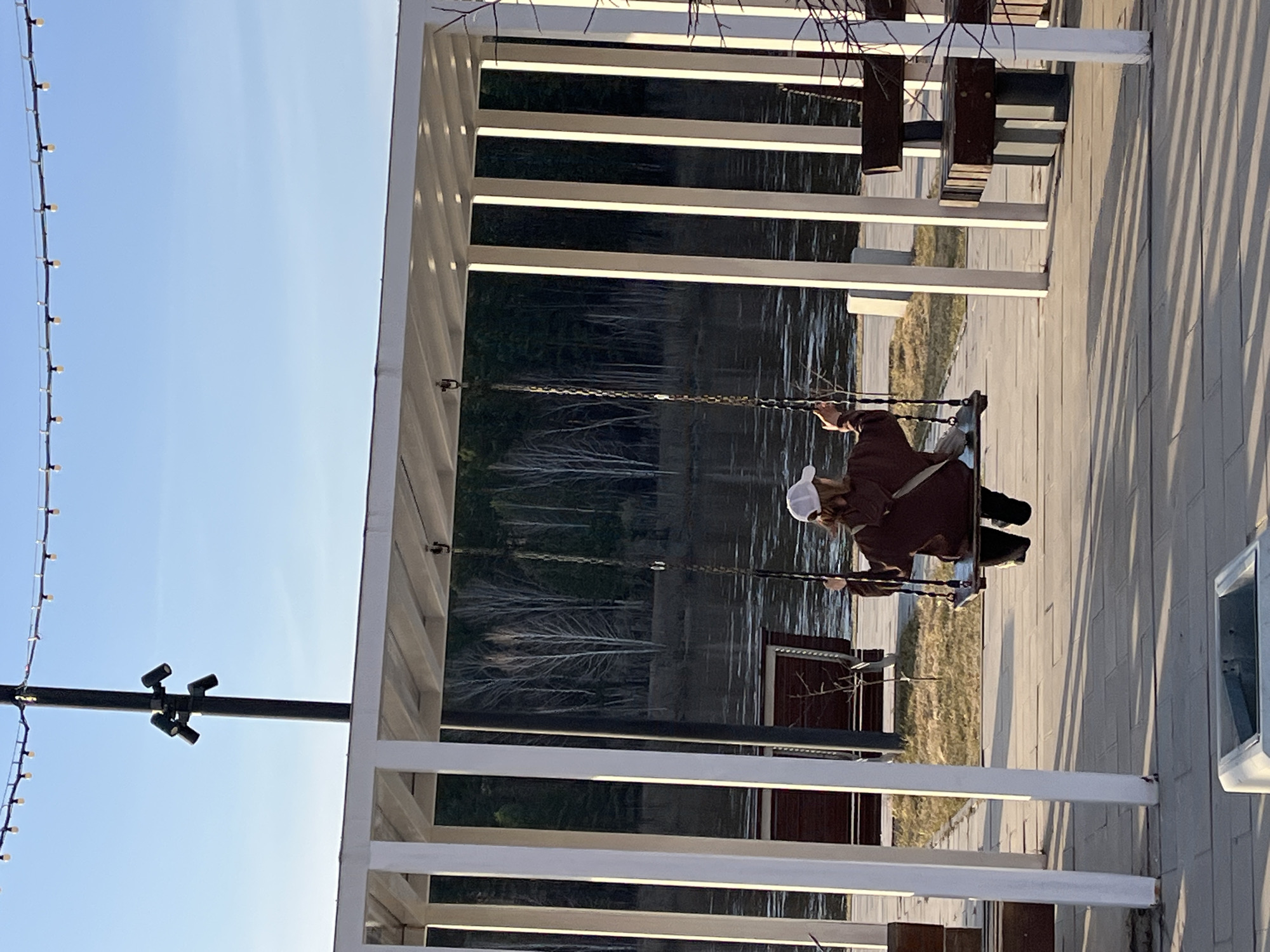 A person is depicted sitting on a swing, looking out over a calm body of water, likely a river or lake, in Zelenogorsk, Russia. The individual is seen from behind, wearing a dark brown coat or jacket, dark trousers, and a white baseball cap. Their posture suggests a moment of quiet contemplation or relaxation.
The swing is suspended by chains from a modern, white pergola structure that features multiple horizontal beams. These beams cast long, distinct shadows onto the light-colored paved ground to the right of the person, indicating that the sun is low in the sky, characteristic of late afternoon or early evening.
Beyond the swing and pergola, a serene expanse of water reflects the soft light. The distant shoreline is visible, featuring sparse, possibly deciduous trees, suggesting a season when leaves have fallen. To the far left of the frame, a string of decorative lights with small, glowing bulbs hangs vertically against a clear, light blue sky. Below these lights, a black pole supports a cluster of small spotlights, further suggesting an illuminated public space. The scene conveys a peaceful atmosphere in an inviting outdoor setting designed for leisure. There is no visible text in the image.