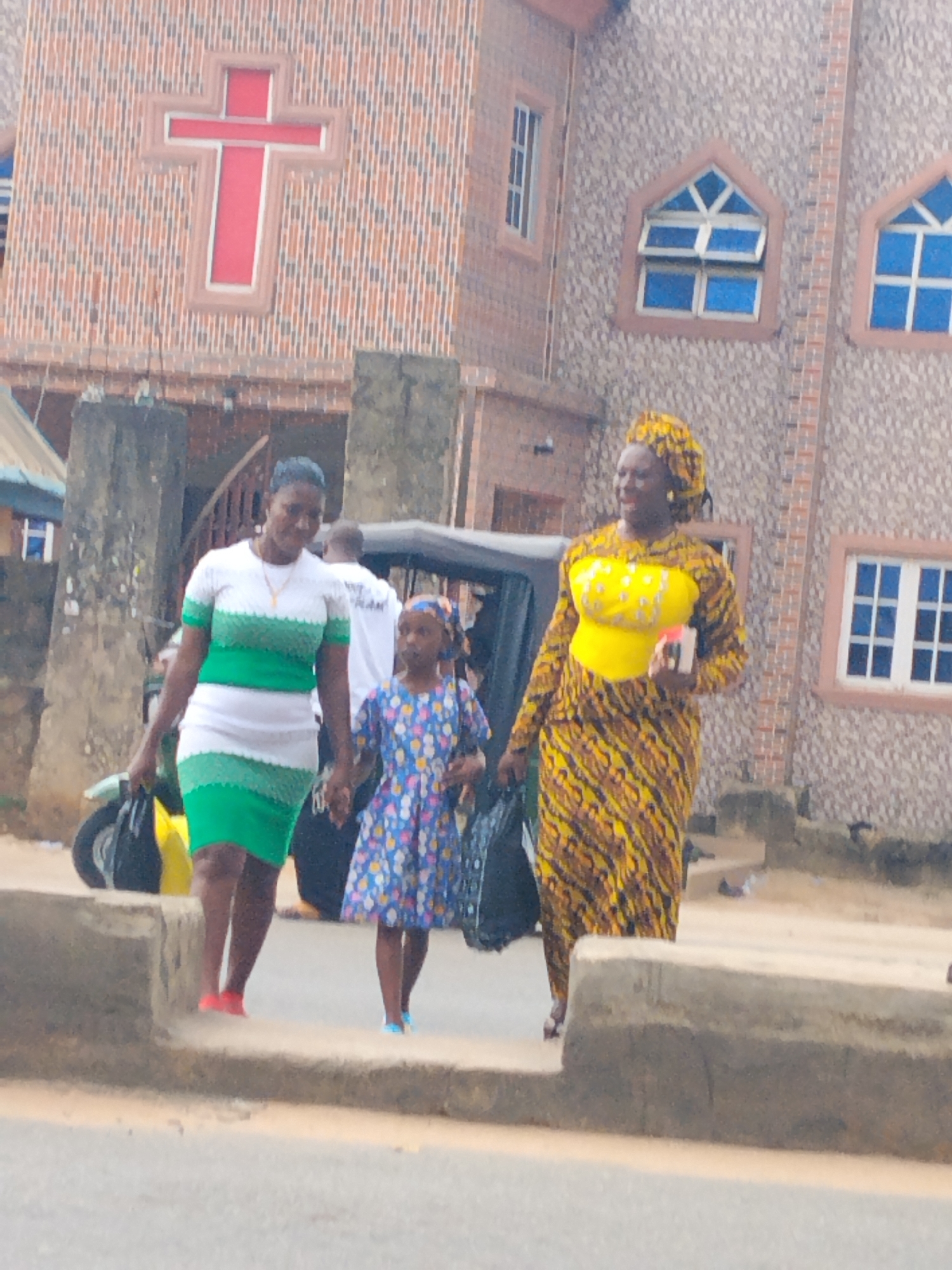 Three people walk on a sidewalk in front of a brick building. The building has a large red cross on its facade and several windows with blue panes. The person on the left is wearing a white and green dress and red shoes. The child in the middle wears a blue dress with a floral pattern and blue shoes. The person on the right is wearing a yellow and brown patterned outfit and a yellow headscarf. A motorcycle and a small vehicle are parked nearby. The city is Aba, Nigeria.