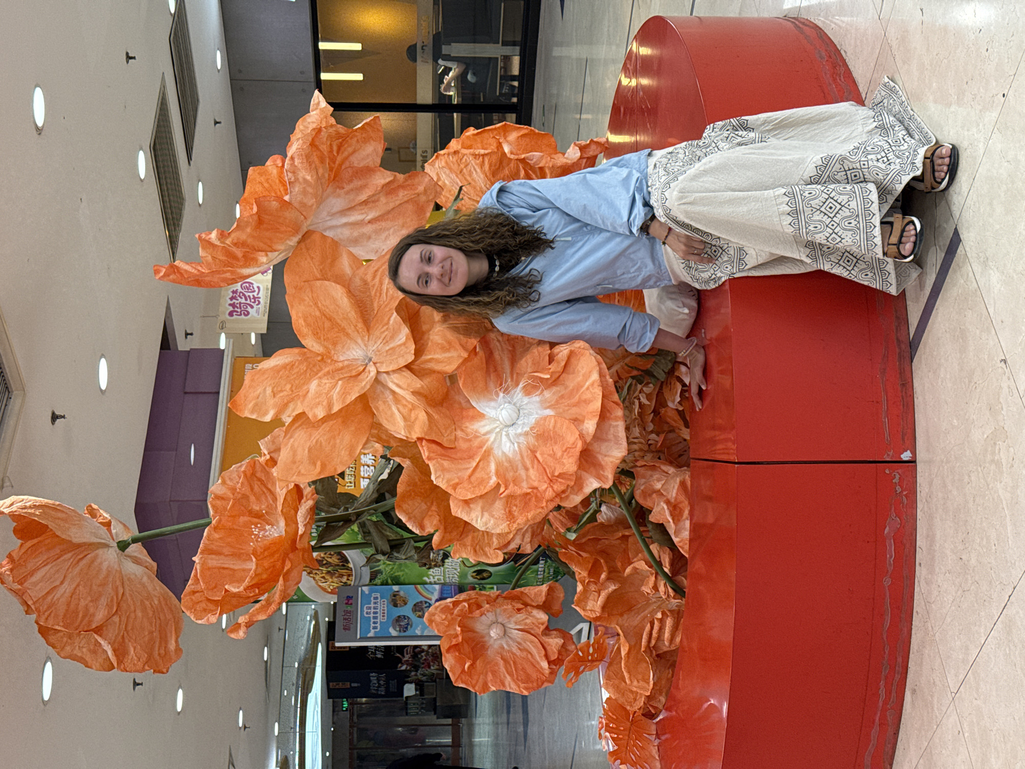 A young woman with curly brown hair smiles while posing on an orange, cylindrical bench surrounded by large, artificial orange flowers. She wears a light blue long-sleeved shirt, a long white skirt with patterned trim, and open-toed sandals. The bench appears to be part of a decorative installation. The location appears to be a shopping mall. Display windows are visible in the background. There is what appears to be Chinese writing on a sign in the background, possibly written in simplified characters. Based on the writing on the sign, the shopping mall may be located in the city of Dongcheng District, China.