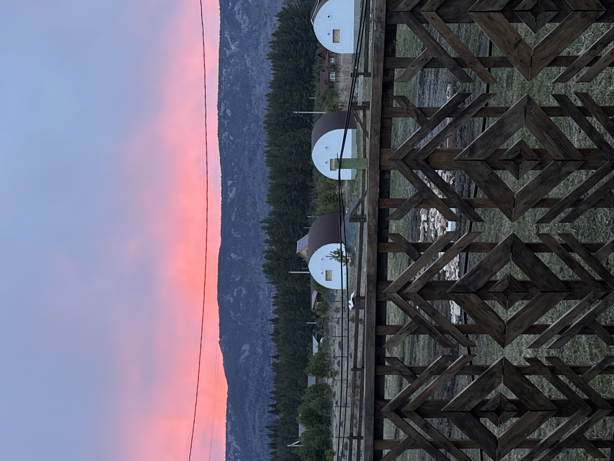 The image captures a picturesque landscape scene, likely at either sunrise or sunset, given the vivid and gradient colors in the sky. No people are visible in the frame.

In the immediate foreground, a striking wooden fence or barrier is present, characterized by an intricate geometric pattern made of dark wooden planks. The design features interconnected diamond and zigzag shapes, giving it a rustic or possibly indigenous aesthetic.

Beyond the fence, the middle ground reveals a grassy field where several unique, dome-shaped structures are situated. These small buildings appear to be modern glamping pods or dwellings, featuring white or light-colored facades and darker, rounded tops. At least one of these structures clearly shows a small, rectangular window. A thin, dark line, possibly a power cable or wire, stretches horizontally across this section of the image. Behind these structures and the grassy area, a dense line of dark green evergreen trees, likely conifers, forms a natural barrier.

In the background, a majestic mountain range rises, its peaks and slopes appearing dark and rugged against the sky. The sky itself is a notable feature, transitioning from a soft, light blue at the top to a vibrant, warm pink and orange glow closer to the mountain horizon, indicative of twilight hours.

The scene conveys a sense of tranquility and natural beauty, combining man-made structures with expansive natural elements. There is no discernible activity taking place, focusing instead on the serene environment. No visible text is present in the image. The location context provided is Tyulyuk, Russia, which aligns with the mountainous and forested landscape depicted.