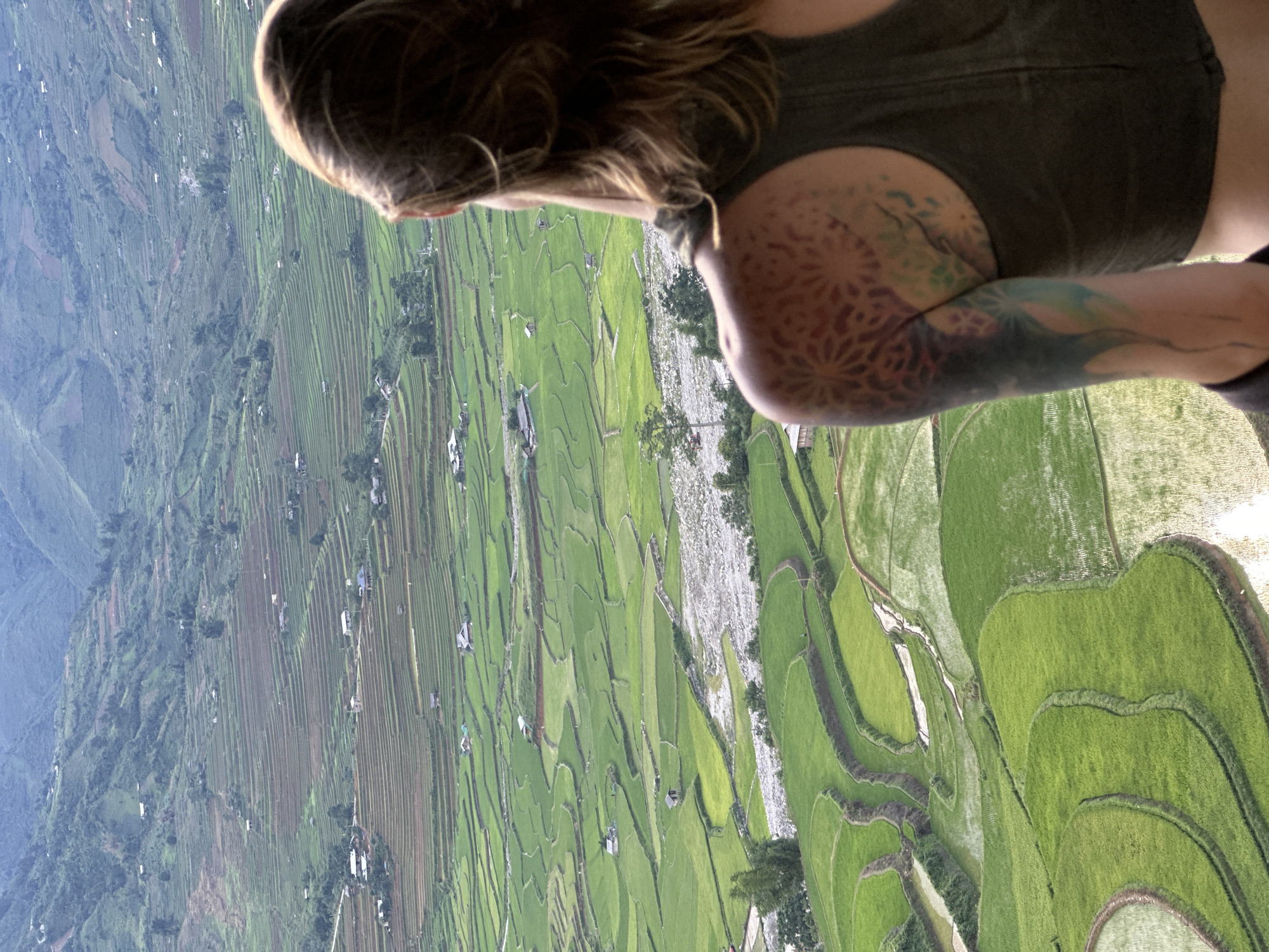 An individual is depicted from a rear-over-the-shoulder perspective, observing a vast terraced landscape in Tú Lệ, Vietnam. The person has medium-length brown hair and wears a dark green sleeveless top. A patterned tattoo, featuring a geometric, mandala-like design in reddish-orange tones, is visible on their left shoulder. Below it, on the upper left arm, is another tattoo displaying abstract forms with blue, green, and yellow hues. The expansive valley below is characterized by numerous vibrant green rice terraces, some partially flooded, conforming to the undulating terrain. A light grey, rocky riverbed, containing visible water, courses through the midground. Scattered throughout the rice fields and on the lower slopes of the hills are multiple small, simple structures, identified as dwellings. The distant background consists of densely vegetated mountains beneath a hazy, light blue sky.