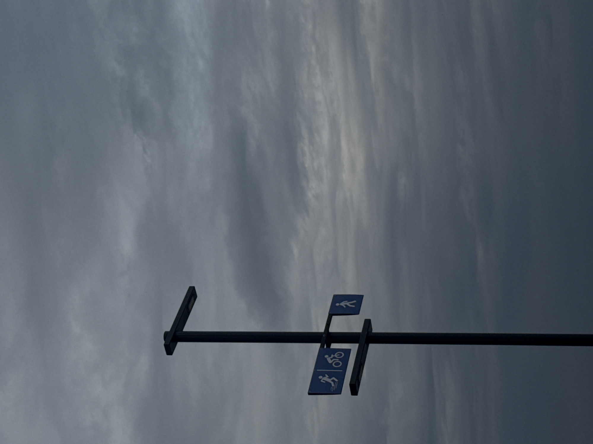 The image captures a minimalist scene of a dark, horizontal pole set against an overcast sky. Three blue rectangular signs with white pictograms are attached to the pole, indicating different types of designated paths. From top to bottom (as oriented in the original image), the signs depict a person walking (pedestrian), a bicycle (cyclist), and a person running or jogging (runner).
The scene is dominated by a cloudy sky, displaying various shades of grey. Lighter, streaky clouds are visible against darker, more uniform patches, suggesting a heavily overcast day with subdued lighting. There is no visible ground or other objects, focusing entirely on the pole, signs, and sky. The lack of direct sunlight indicates either an early morning, late afternoon, or simply a very cloudy day. The setting is likely an outdoor public space, such as a park, shared pathway, or recreational area in Chaikovsky, Russia, where these signs would direct users. No specific activity is taking place within the frame, but the signs themselves imply the presence or allowance of these activities. There is no visible text, only the internationally recognized pictograms.