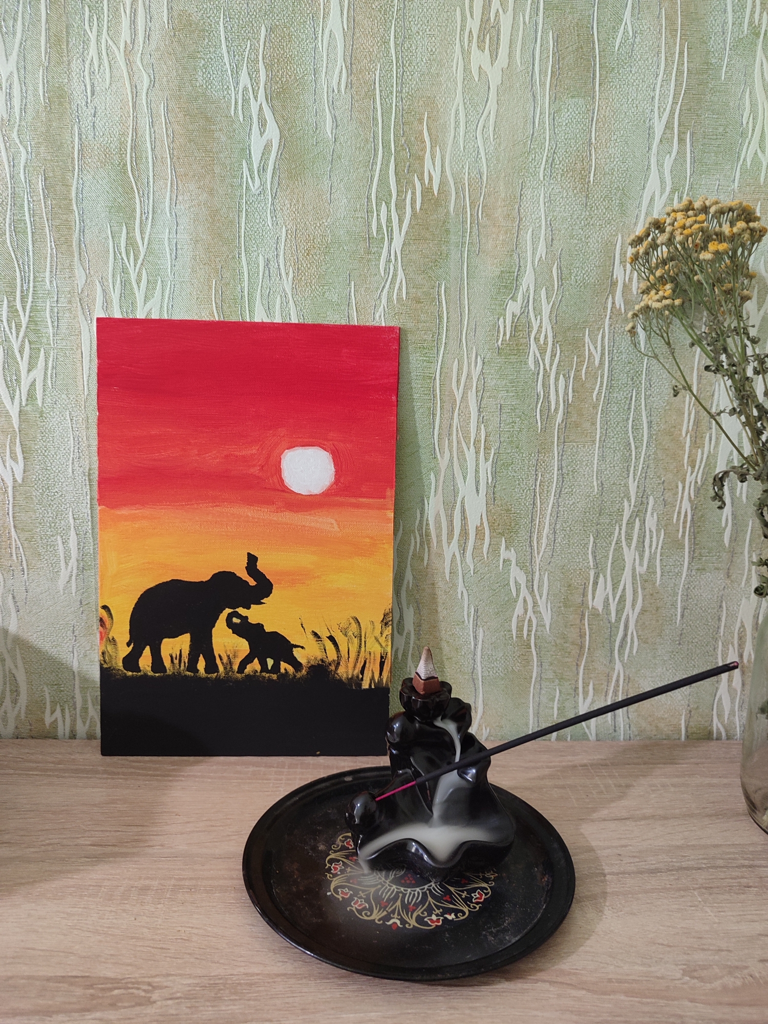 The image depicts a still life composition placed on a wooden shelf against a patterned wallpaper. A rectangular painting of elephants silhouetted against a sunset sky is positioned to the left. In the foreground, a black ceramic incense burner is emitting smoke from a lit incense stick, which is resting on a decorative black plate. To the right of the painting and incense burner, a small bouquet of dried yellow flowers in a clear glass vase is visible. The overall scene appears to be indoors, likely on a shelf or tabletop, with the wallpaper serving as the backdrop. There are no people or animals present in the immediate scene other than the painted elephants. The location is specified as Lozuvatka, Ukraine, though no specific landmarks or contextual elements in the image directly indicate this geographical location. The lighting suggests an ambient indoor setting, possibly with natural light from a window out of frame.