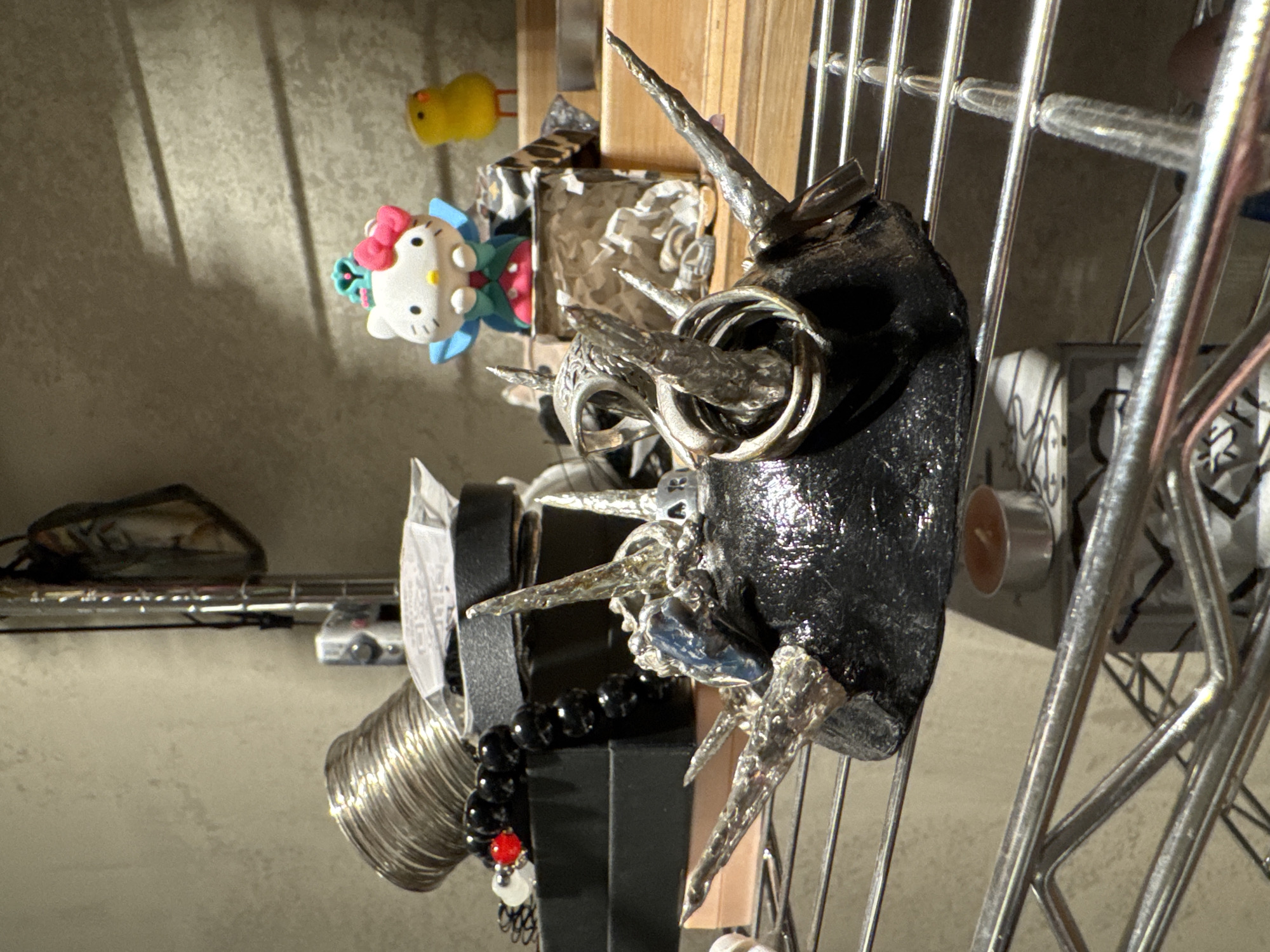 A shelf in a room contains various objects, including a spiked, dark, textured object with rings and a blue gem in the foreground. Behind it are black beaded bracelets with a red bead, a silver coiled object, and a black box. Further back, a figurine of Hello Kitty in a blue and green dress with a pink bow sits on a tiered structure, next to a small yellow chick figurine. A lit tea light candle is visible on a lower shelf. The background appears to be a textured wall. The image is taken from a low angle, looking upwards at the shelf.