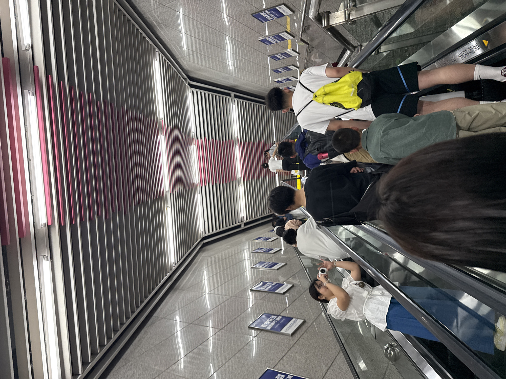 The image shows a group of people riding an escalator in what appears to be a subway station in Guang'an, China. The background features a wall with a decorative design consisting of vertical stripes in shades of gray and pink, interspersed with fluorescent lights. The floor shows signage and the reflections from the ceiling lights. Several people are visible on the escalator, some wearing casual clothing like T-shirts, shorts, and pants. One person is taking a photo with a digital camera. There is an absence of prominent branding or logos.