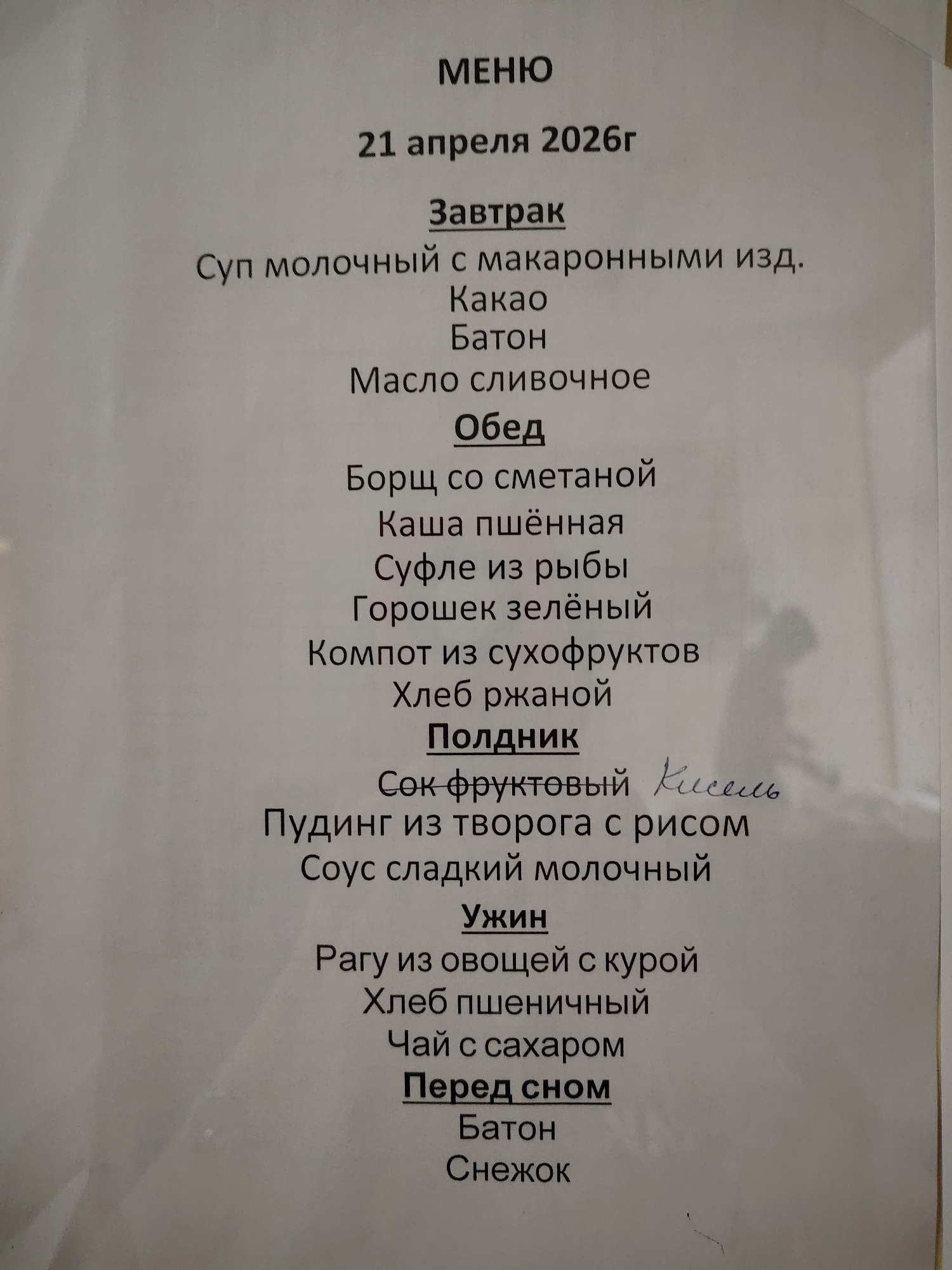 The image displays a close-up of a printed menu from Sarapul, Russia, for April 21, 2026. The menu is presented on a light-colored sheet of paper, possibly slightly creased or uneven, against a neutral background.
The text is predominantly in black, bolded for meal categories. It outlines five meals for the day:
**МЕНЮ (MENU)**
**21 апреля 2026г (April 21, 2026)**
**Завтрак (Breakfast)**
* Суп молочный с макаронными изд. (Milk soup with pasta products)
* Какао (Cocoa)
* Батон (Loaf of white bread)
* Масло сливочное (Butter)
**Обед (Lunch)**
* Борщ со сметаной (Borscht with sour cream)
* Каша пшённая (Millet porridge)
* Суфле из рыбы (Fish soufflé)
* Горошек зелёный (Green peas)
* Компот из сухофруктов (Dried fruit compote)
* Хлеб ржаной (Rye bread)
**Полдник (Afternoon Snack)**
* Сок фруктовый (Fruit juice) - with "Кисель" (Kissel, a thick fruit drink) handwritten next to it.
* Пудинг из творога с рисом (Cottage cheese pudding with rice)
* Соус сладкий молочный (Sweet milk sauce)
**Ужин (Dinner)**
* Рагу из овощей с курой (Vegetable stew with chicken)
* Хлеб пшеничный (Wheat bread)
* Чай с сахаром (Tea with sugar)
**Перед сном (Before Sleep)**
* Батон (Loaf of white bread)
* Снежок (Snezhok, a fermented milk drink)
The lighting is even, suggesting an indoor setting. There are no people or discernible activities, focusing solely on the menu's contents.