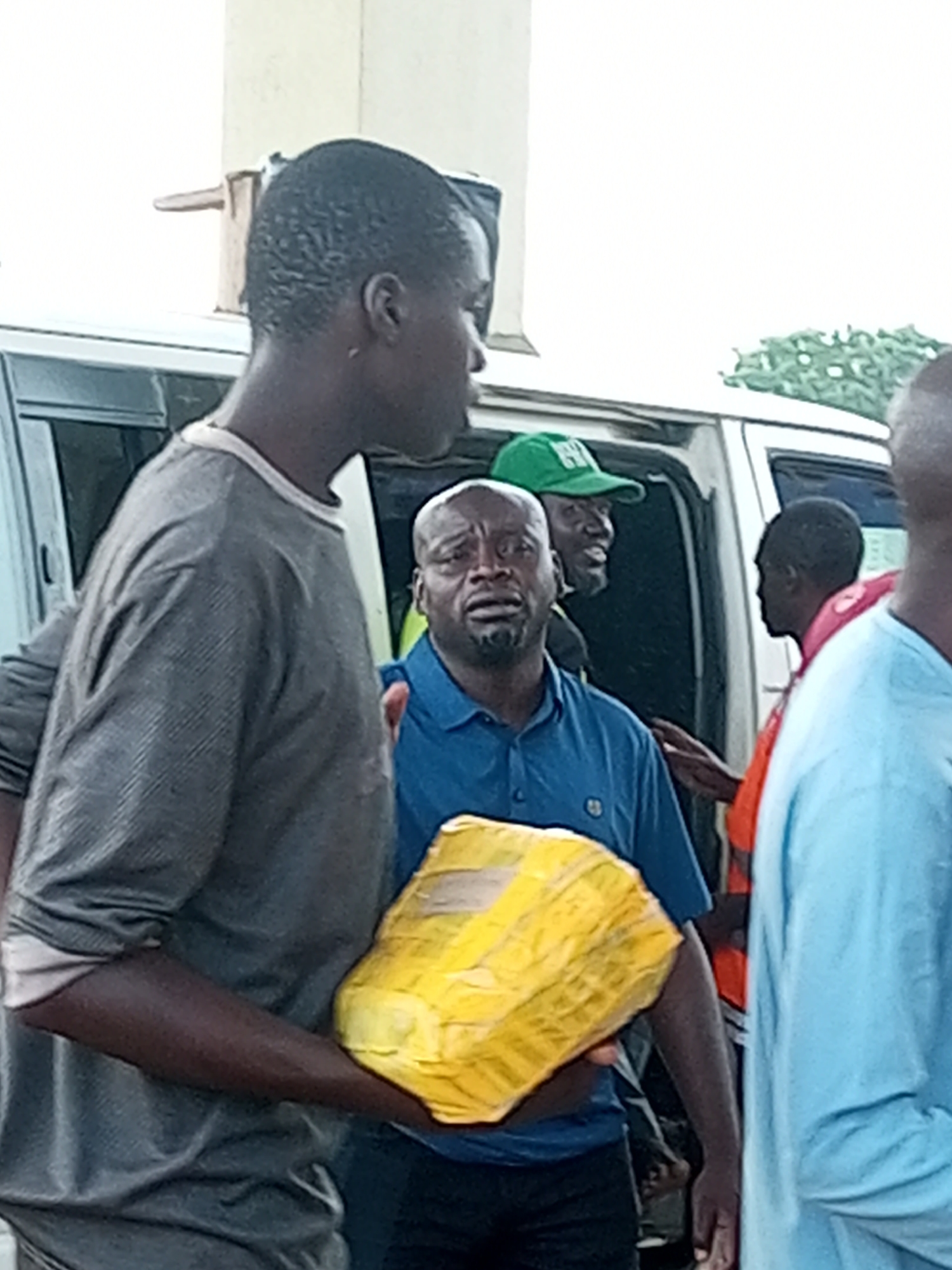 The image depicts a group of people in Gombe, Nigeria, near a white van. In the foreground, a man in a grey long-sleeved shirt is partially visible from the side, with his arm extended forward. He appears to be holding a yellow-wrapped package. Next to him, a man in a blue polo shirt looks distressed, with his mouth open and eyes wide. He is also holding the yellow package. Behind them, a man wearing a green baseball cap with white lettering is visible inside the van, smiling and looking towards the right. Another person in an orange shirt and a person in a light blue shirt are also present. The setting appears to be outdoors, possibly in a street or parking area. The lighting suggests it is daytime, and the overall atmosphere seems tense or dramatic due to the expressions of some individuals and the packaging of the item. No specific text is clearly legible in the image.