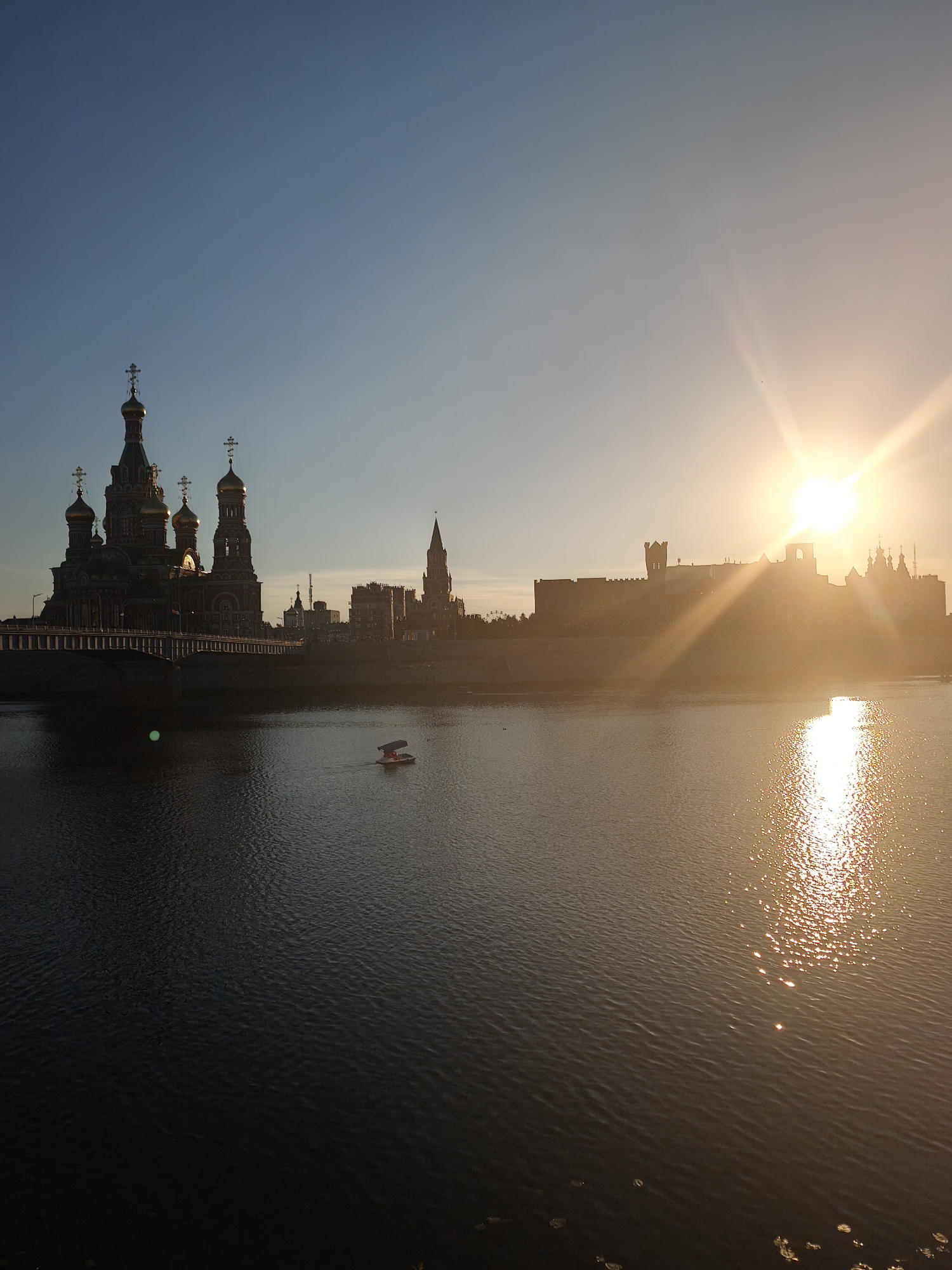 At sunset in Yoshkar-Ola, Russia, the Tsarevokokshaysky Kremlin and the Church of the Holy Archangel Gabriel stand silhouetted against a bright sky. A river flows in the foreground, reflecting the golden light of the setting sun. A small pedal boat with two occupants navigates the water. The overall scene is tranquil and picturesque, capturing the beauty of the city at dusk.
