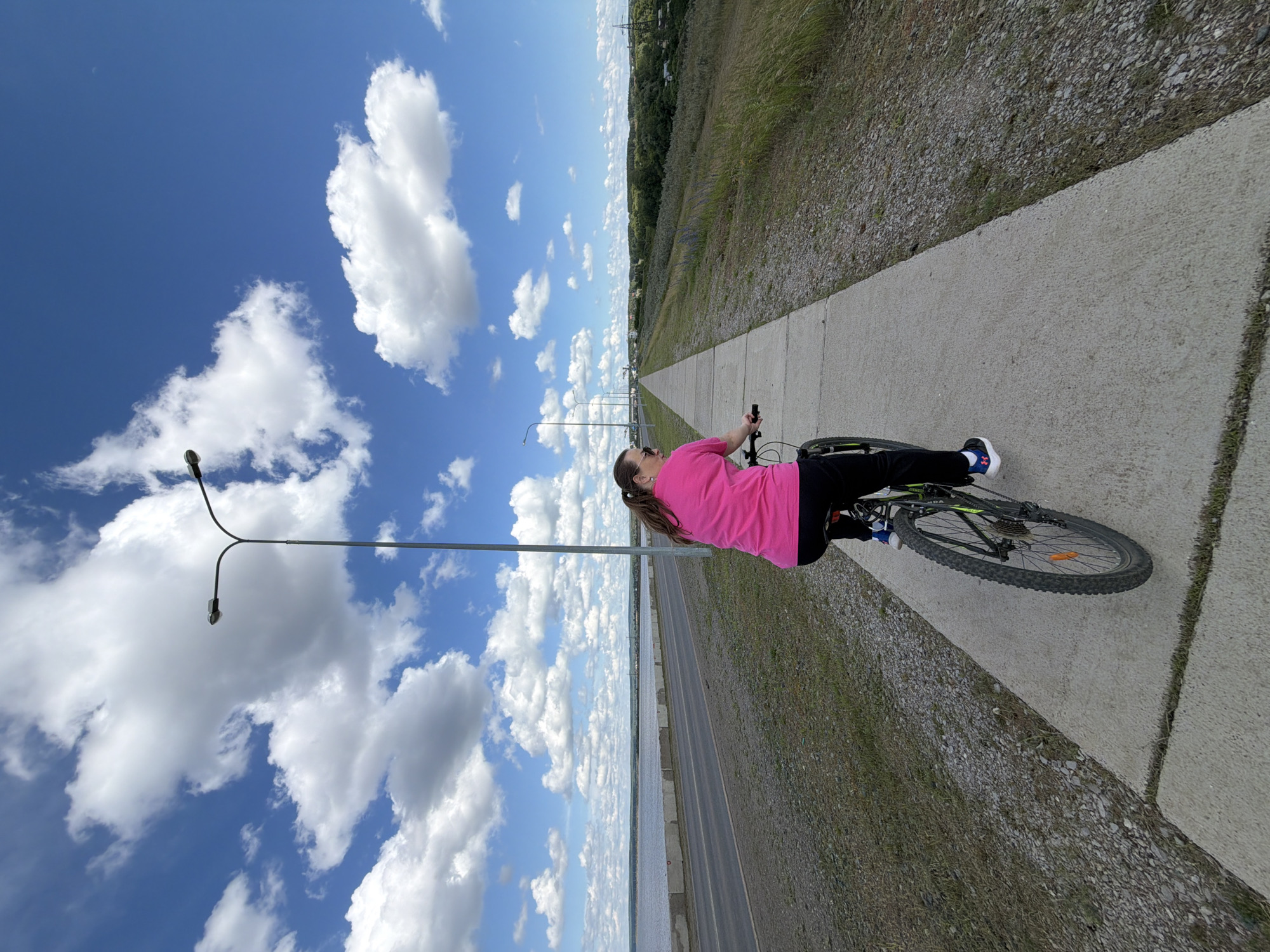 A person rides a bicycle on a paved path next to a road. The person is wearing a bright pink shirt and black pants. The bicycle is dark colored with green accents. In the background, there is a body of water, a road with cars, and a landscape with trees and houses. The sky is blue with large white clouds and there are streetlights along the path. The location is the embankment of the Kama River in Chaikovsky, Russia.