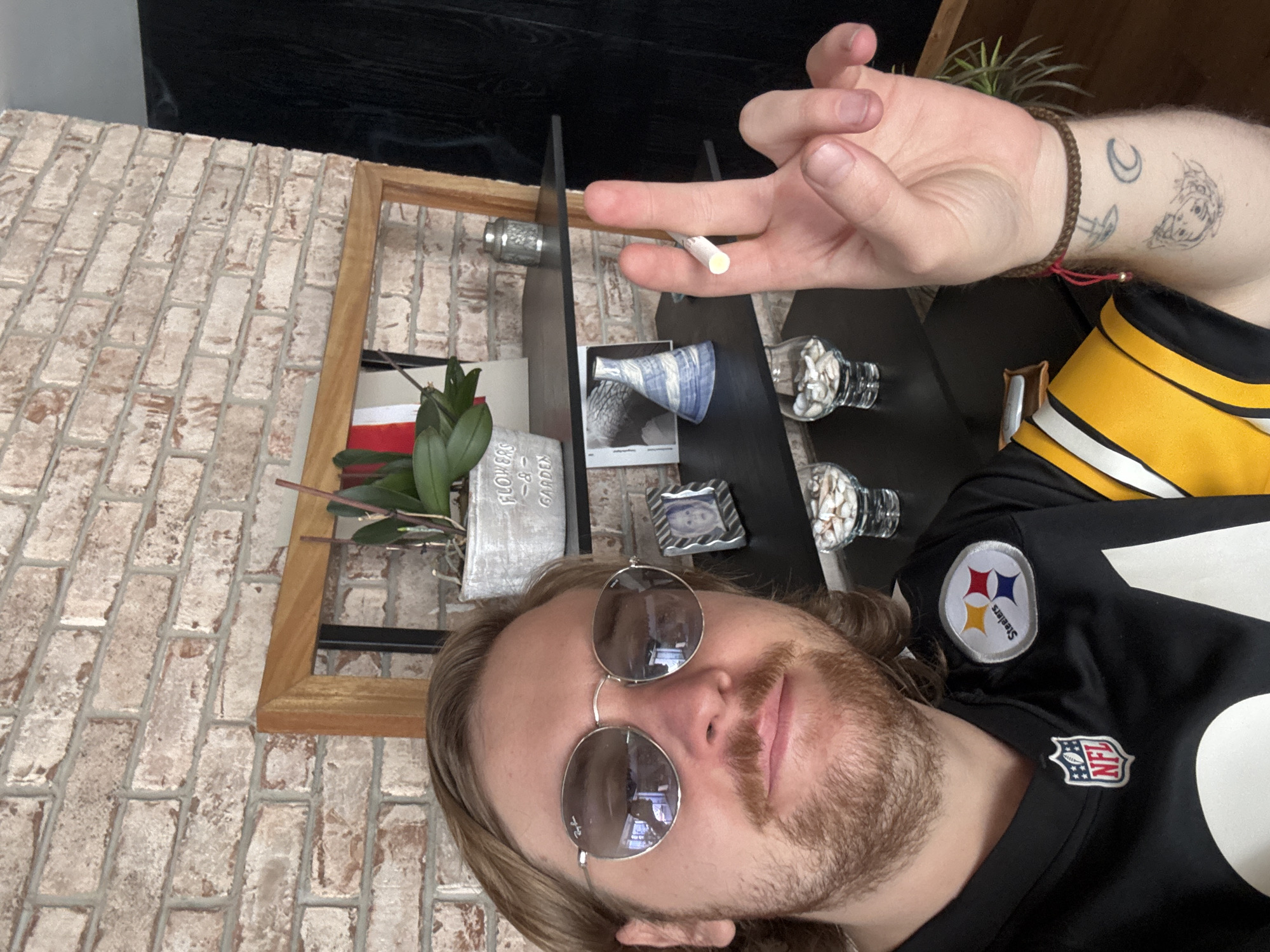 A Caucasian male with long blonde hair, a mustache, and a beard is seen taking a selfie. He is wearing round sunglasses and a Pittsburgh Steelers jersey. He holds a thin, white cylinder object, resembling a cigarette or a joint, between his fingers. His right forearm is visible, adorned with a woven bracelet and tattoos.
Behind him, there is a shelf displaying decorative items, including a potted plant labeled "Flowers & Garden," small decorative vases and containers. The shelf is positioned against a brick wall.
The image provides no information to ascertain the location in Huixquilucan de Degollado, Mexico.