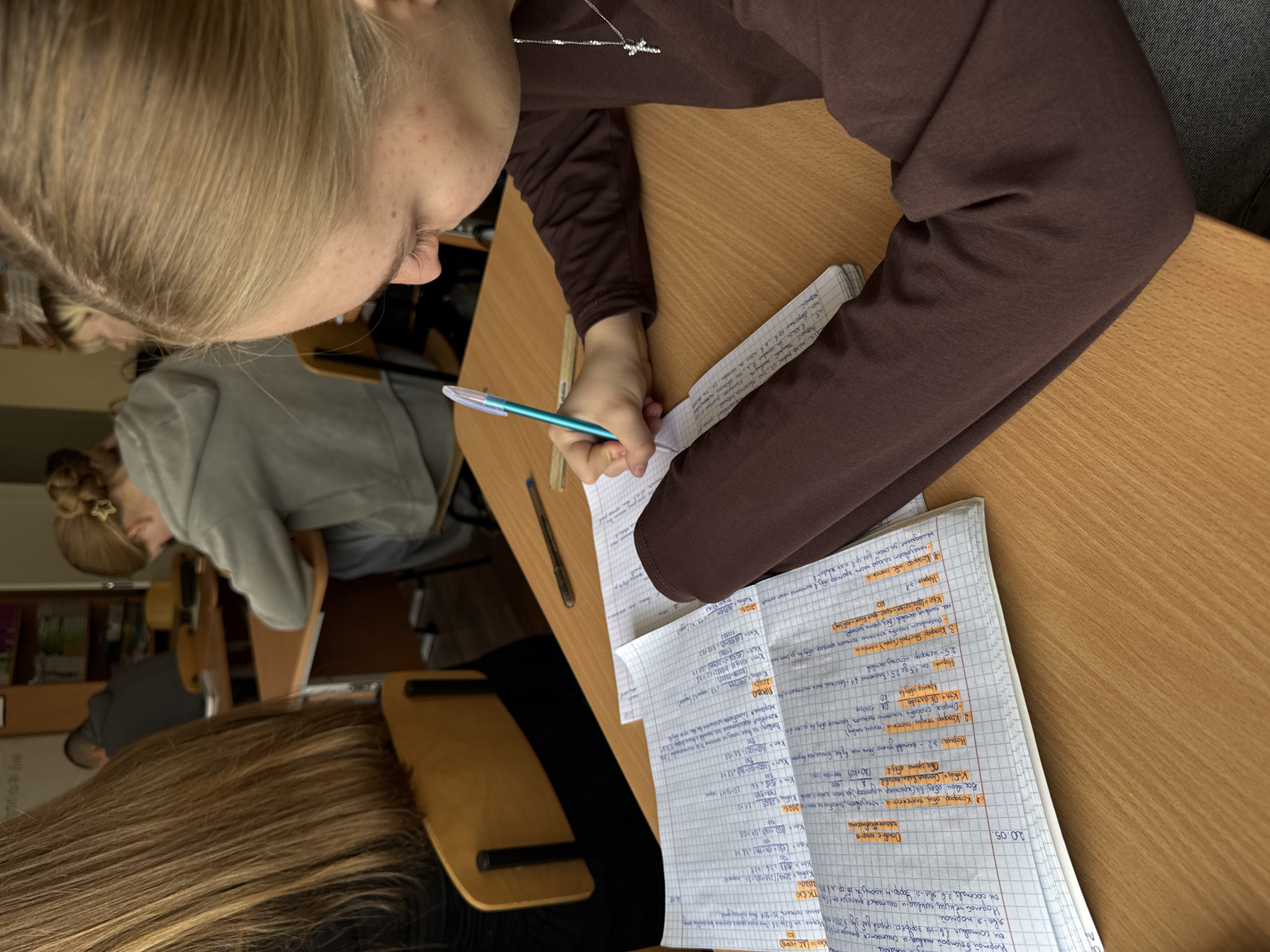 This photograph captures a scene within a classroom, likely in Perm, Russia. A student with blonde hair, wearing a brown long-sleeved shirt, is actively writing in a notebook filled with handwritten notes, possibly related to finance, as indicated by calculations and terms like "платежест" (solvency) and "активов" (assets). The notebook features lines and seems to be partially filled with formulas and annotations, some highlighted in orange. A pen is held in the student's hand, poised above the page.
The desk is a light-colored, possibly wooden surface. Other students are visible in the background, one with blonde hair pulled up into a bun wearing a gray sweatshirt. In the background are bookshelves, and other desks. The image is taken from an overhead perspective, focusing on the immediate area of the student and their work. The overall environment suggests an educational setting, possibly a university or advanced secondary school.