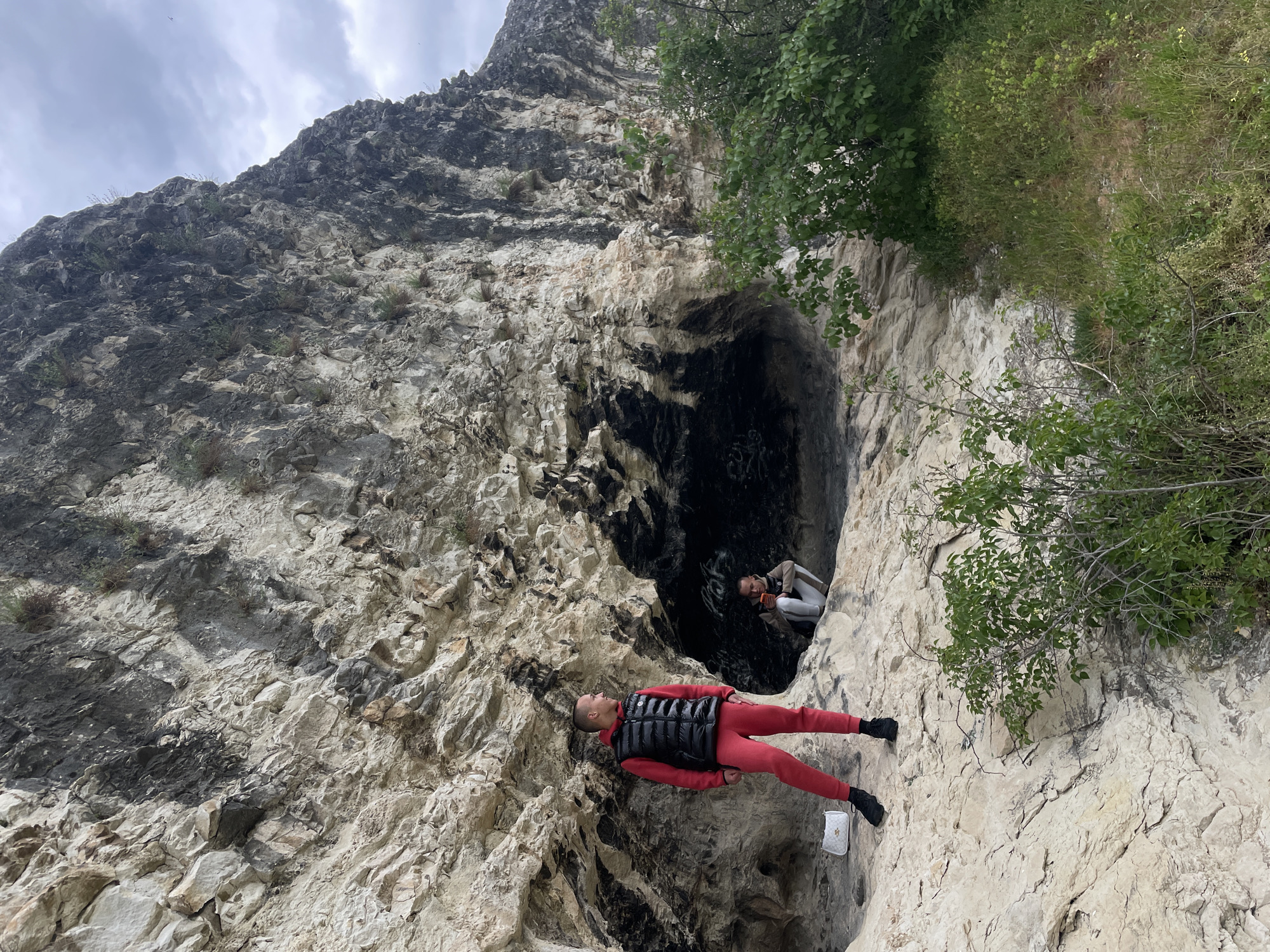 The image displays two individuals interacting with a rugged, textured rock face featuring a prominent dark cave opening. The photograph is oriented with a 90-degree clockwise rotation, making the rock face appear as a steep, almost horizontal plane across the frame.
In the foreground, one person is lying horizontally on their back on a rock ledge. They are dressed in a red tracksuit, a black puffer vest, and black shoes. A small white handbag with a distinctive patterned design, including what appears to be the "LV" monogram, is placed near their feet.
Further into the scene, within the dark entrance of the cave, a second person is visible. They are wearing light-colored clothing and holding an orange object, possibly a mobile phone, while looking outwards.
The setting is an outdoor, mountainous or cliff-side environment with irregular rock formations ranging from light gray to darker, shadowed patches. Sparse green foliage and bushes are scattered across the rock and along the top edge of the cliff. The sky, visible in the upper-left portion of the rotated image, is overcast with clouds. The scene appears to be a deliberate, posed photograph taken in a scenic, rugged location during daytime.