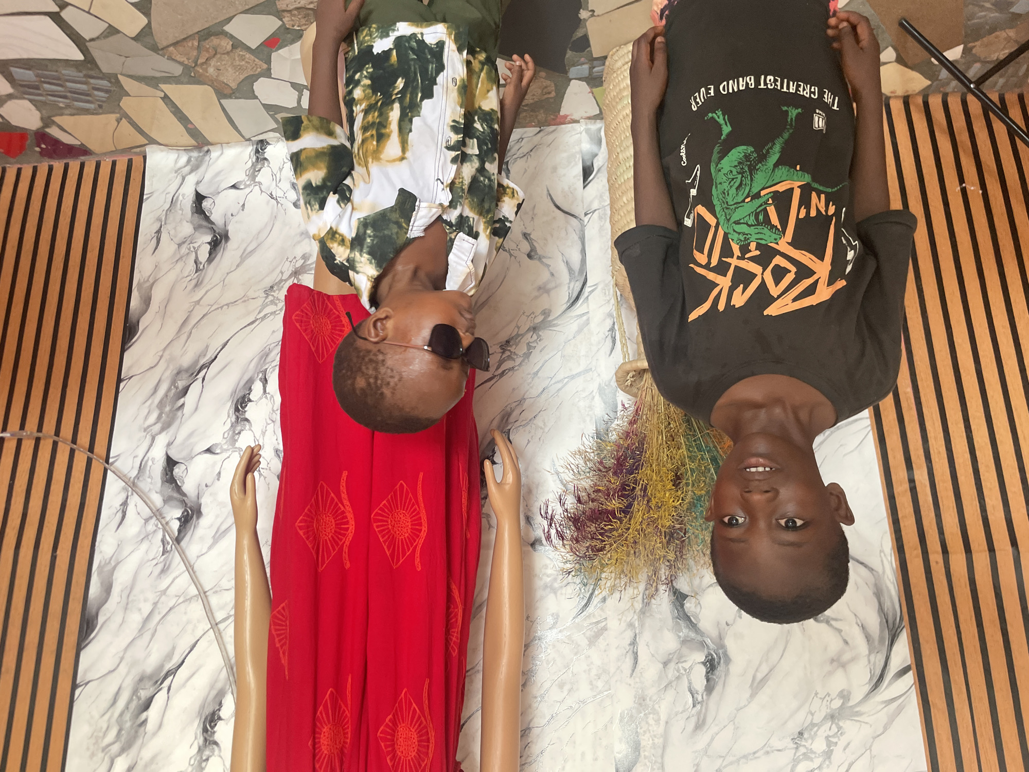 Two people are on the floor. One person wears sunglasses, a white and green button-down shirt, and a red garment. The other person wears a black t-shirt. The scene includes mannequin arms, a bag, and dried plant material. The location is Badagry, Nigeria.